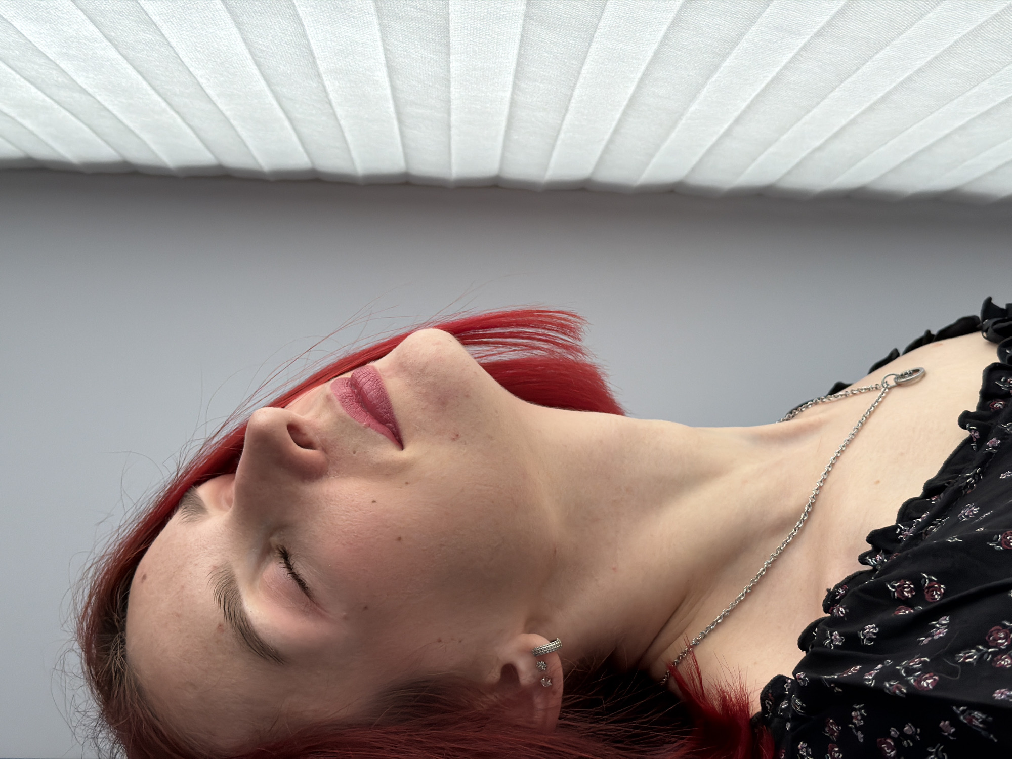 A fair-skinned female with red hair is lying down in a brightly lit interior. She is wearing a black floral-patterned top, a silver necklace, and earrings. Her eyes are closed, and she is wearing lipstick. Above her is a white, folded, textured surface, likely a window covering. The background is a neutral gray. The location is unknown, and there is no visible connection to the city of Chaikovsky, Russia.
