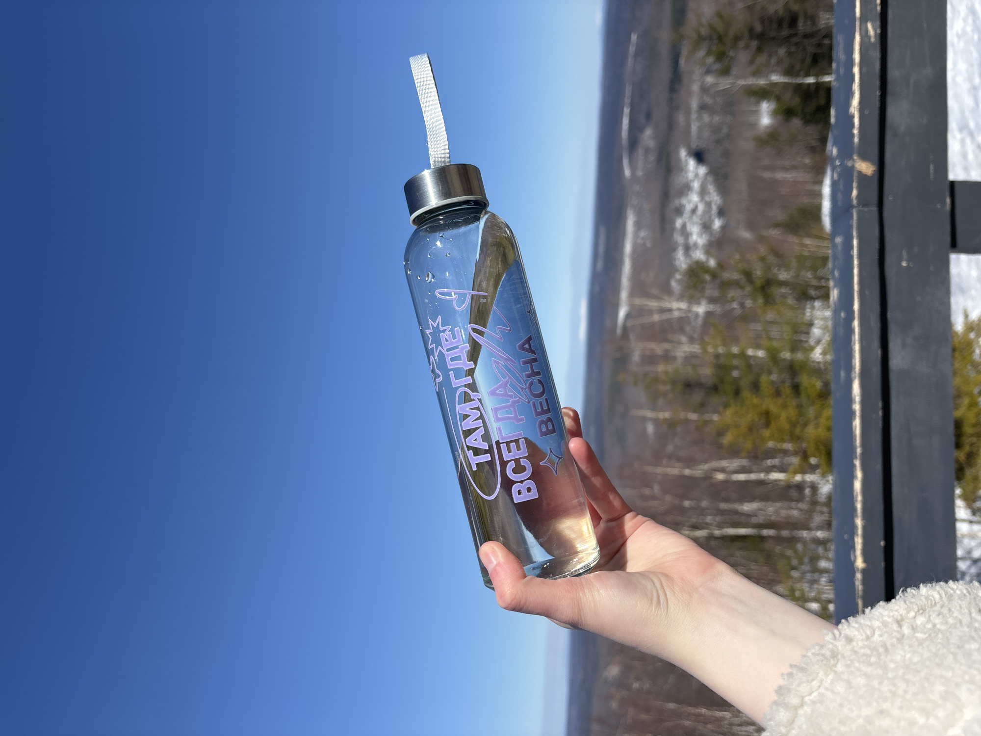 A clear, cylindrical water bottle with a silver lid and white strap is held up against a bright blue sky. The bottle is decorated with purple text that reads "Там где всегда весна" and stars. The hand holding the bottle is fair-skinned. In the background, there is a blurry landscape with trees and patches of snow. A black wooden structure is visible to the right of the bottle. The location is identified as Gubakha, Russia.