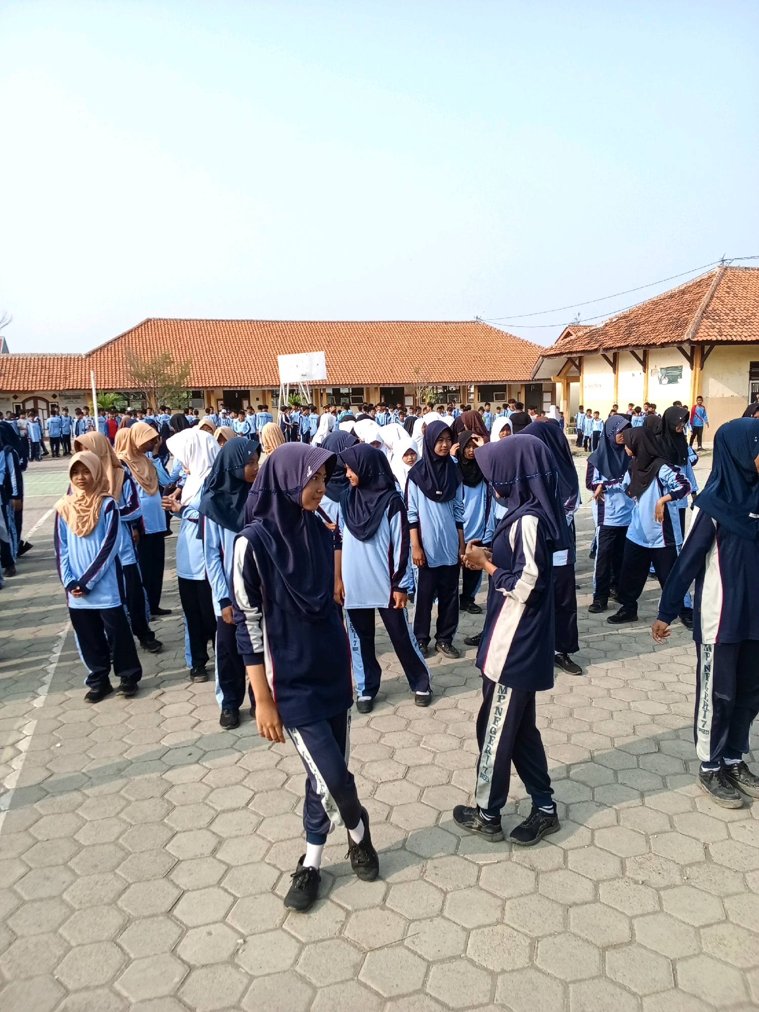The image depicts an outdoor scene at a school in Tengki, Indonesia. A group of students, predominantly female and dressed in matching navy blue and light blue uniforms with headscarves, are gathered in an open courtyard paved with hexagonal concrete tiles. The students appear to be engaged in some form of assembly or activity. In the background, there are buildings with orange tiled roofs and a basketball hoop. The sky is clear and blue, suggesting a sunny day. The overall impression is one of an organized school gathering.