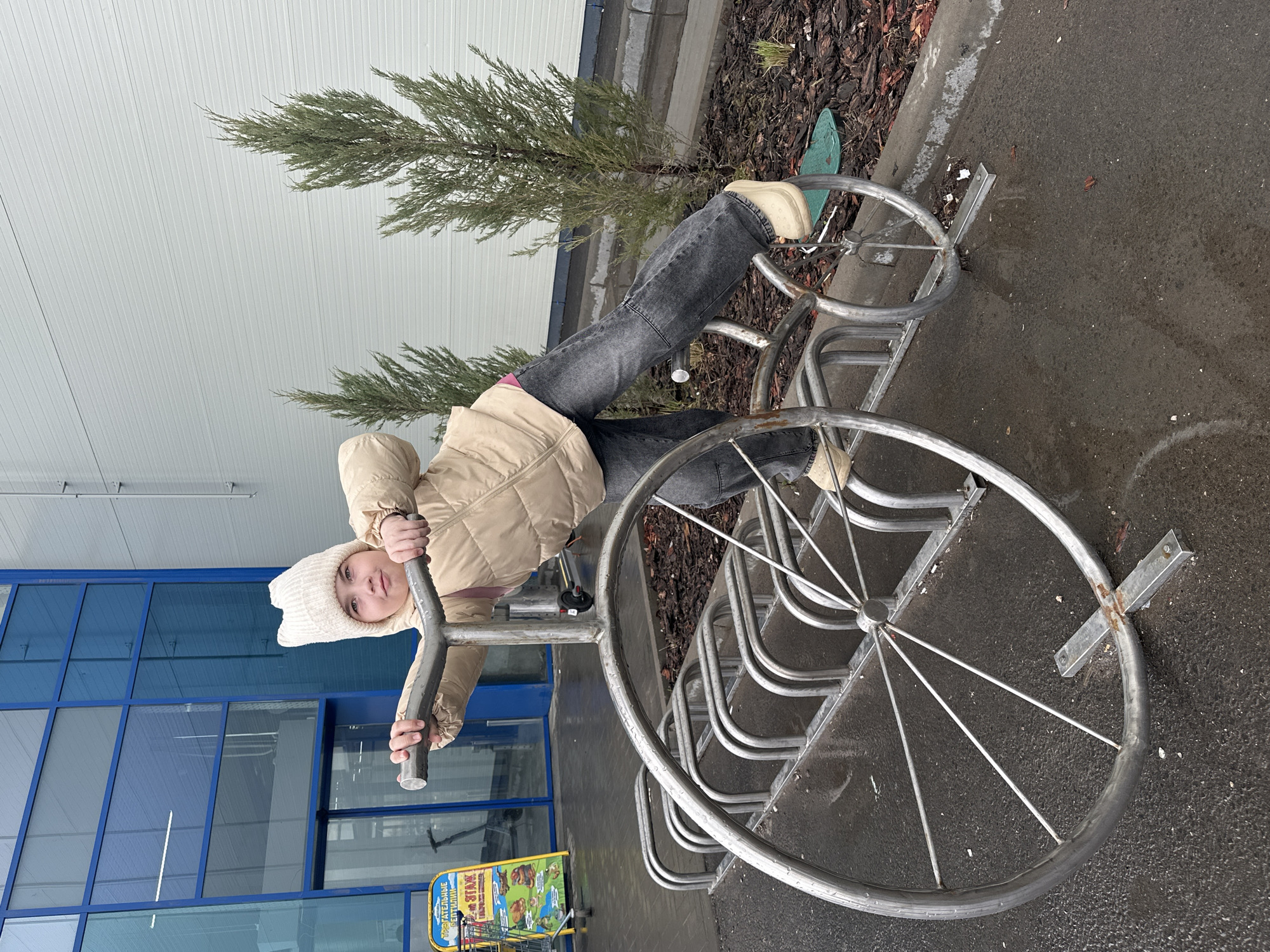 A young girl is positioned on a metal bicycle rack in what appears to be an urban environment. She is wearing a beige jacket, grey jeans, and a white hat. The bicycle rack has large, decorative wheels, and is situated on a wet, paved surface with a tree and building in the background. There is a shopping cart with a promotional sign next to the rack. The location is Izhevsk, Russia.