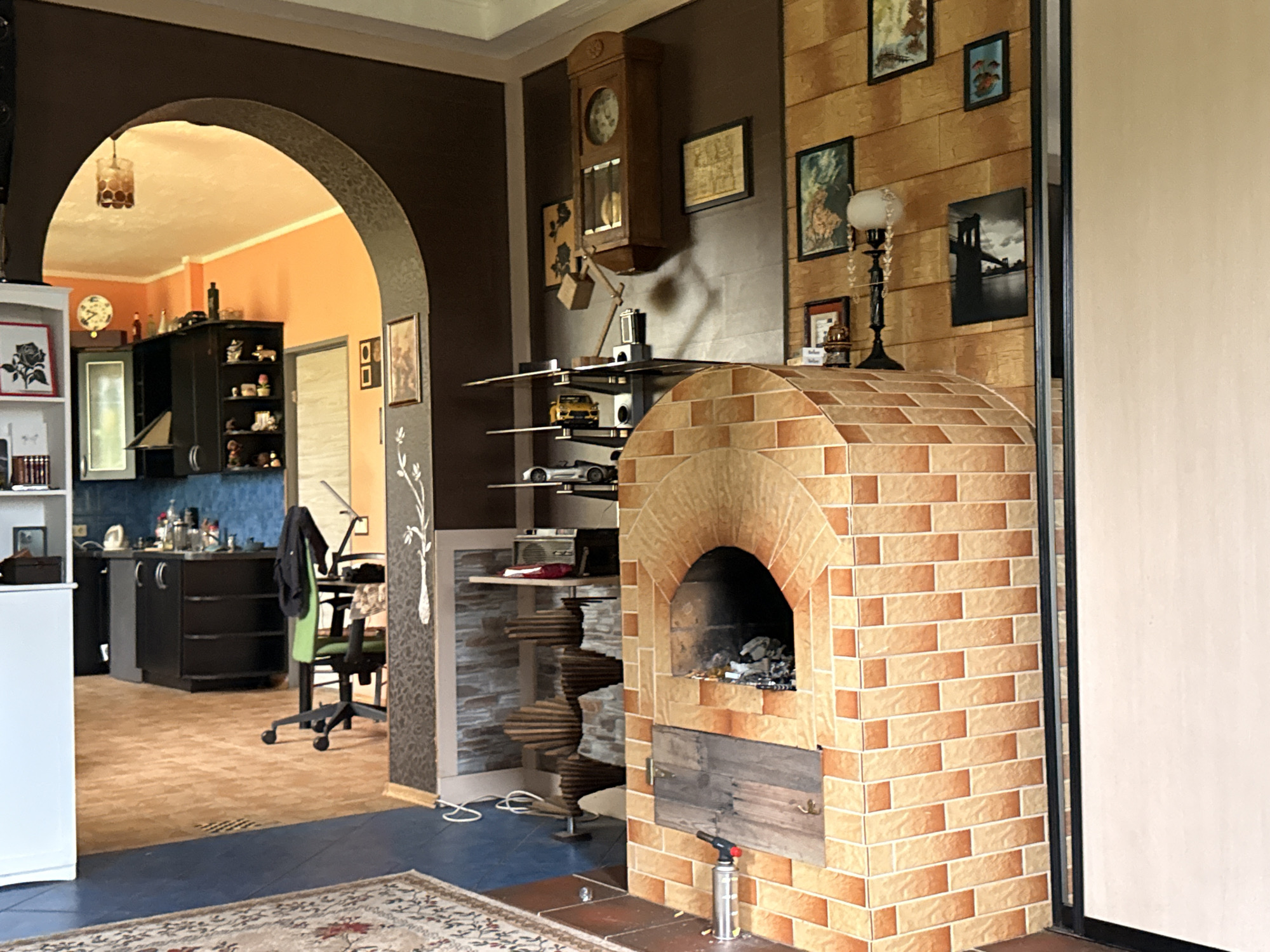 The image depicts an indoor scene, likely a living room or dining area, with an adjoining kitchen visible through an arched doorway. The room features a prominent brick oven with an arched opening, made of light orange and cream-colored bricks. A metal propane torch rests on the floor in front of the oven. To the left of the oven, there is a dark brown wall with several framed pictures and a vintage wooden clock hanging prominently. Below the clock, a multi-tiered shelf holds decorative items and model cars. Further left, through the archway, a kitchen area is visible with dark cabinets, a blue tiled backsplash, and kitchen appliances. A green office chair is positioned in the kitchen area. The floor is a combination of dark blue and light brown wood-like material, with a patterned rug in the foreground. The overall impression is a cozy, eclectic interior. The provided information indicates this scene is located in Galinė, Lithuania.