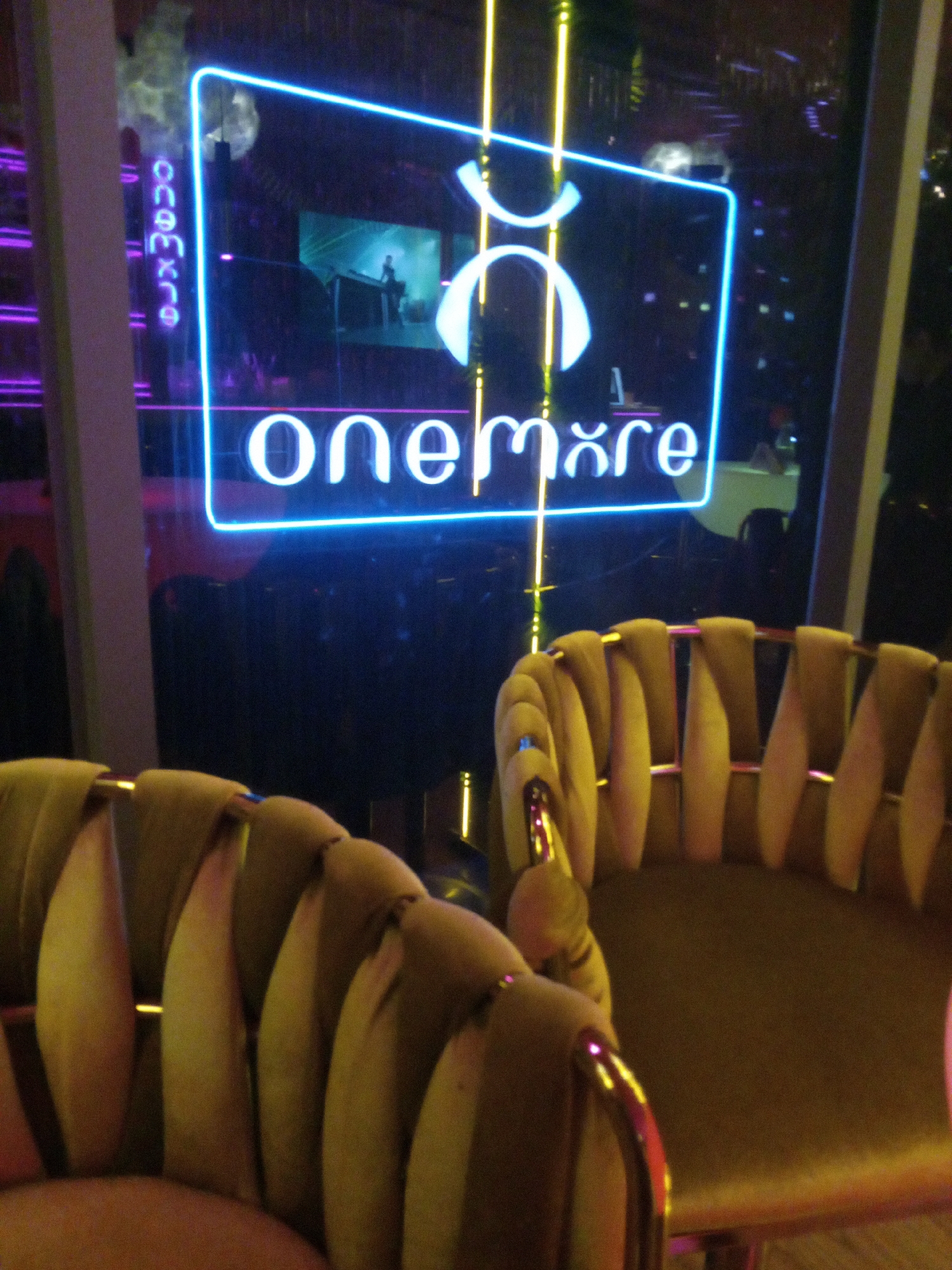 The image shows a bar or lounge setting in Nha Trang, Vietnam. Two mustard-colored velvet chairs with gold metal frames are in the foreground. In the background, a neon sign with the words "onemore" is visible, reflecting in a glass window. The sign also contains a stylized symbol above the text. The location is suggested by the Vietnamese lettering visible vertically on the left of the neon sign. The interior has low lighting, with additional light sources visible as vertical yellow lines in the background. A video screen is visible behind the neon sign.