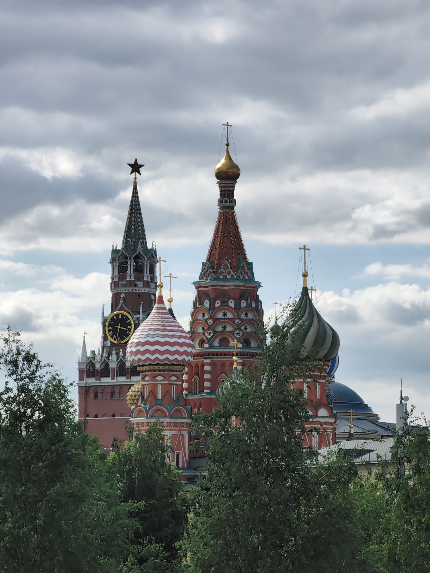 The image features the iconic architecture of Moscow, Russia, specifically Saint Basil's Cathedral and the Spasskaya Tower of the Kremlin. Saint Basil's Cathedral is characterized by its multiple, brightly colored, and uniquely shaped domes, with patterns including stripes and geometric designs in red, white, green, and blue. The Spasskaya Tower is visible to the left of the cathedral, featuring a green spire topped with a red star, and a large clock face. The buildings are set against a cloudy sky with patches of blue. The lower portion of the image is filled with green trees, partially obscuring the base of the buildings. Other buildings are visible behind the trees, including other parts of the Kremlin complex.