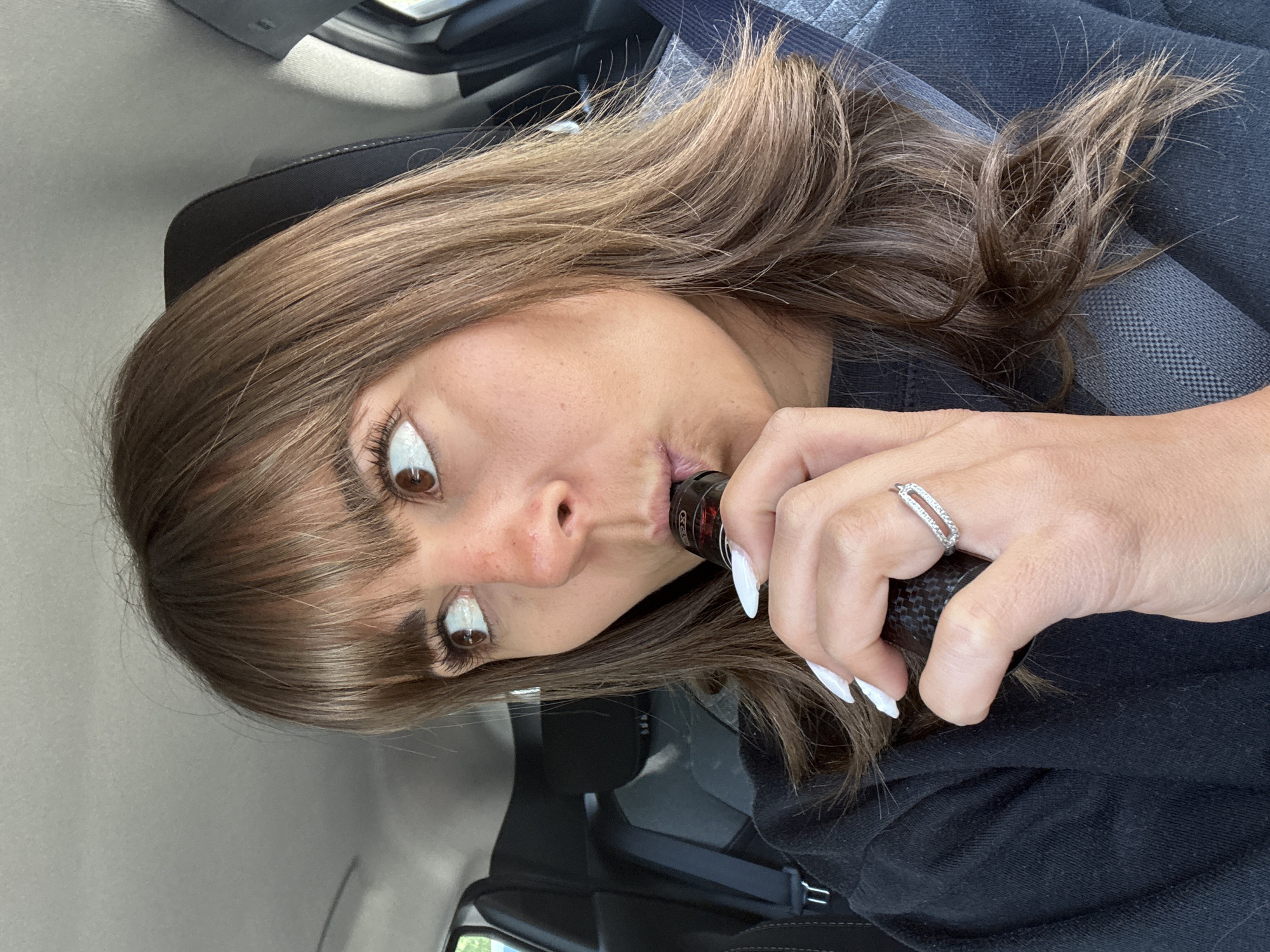 A woman is in the passenger seat of a vehicle. She has brown hair, bangs, and brown eyes. She is holding a vaping device to her mouth. She is wearing a black shirt. Her left hand has long white fingernails, and she is wearing a ring with small gems. The background includes the interior of the car, including the dashboard, seat, and seatbelt. The location is not evident from the image, so the city of Orange, France, cannot be confirmed.