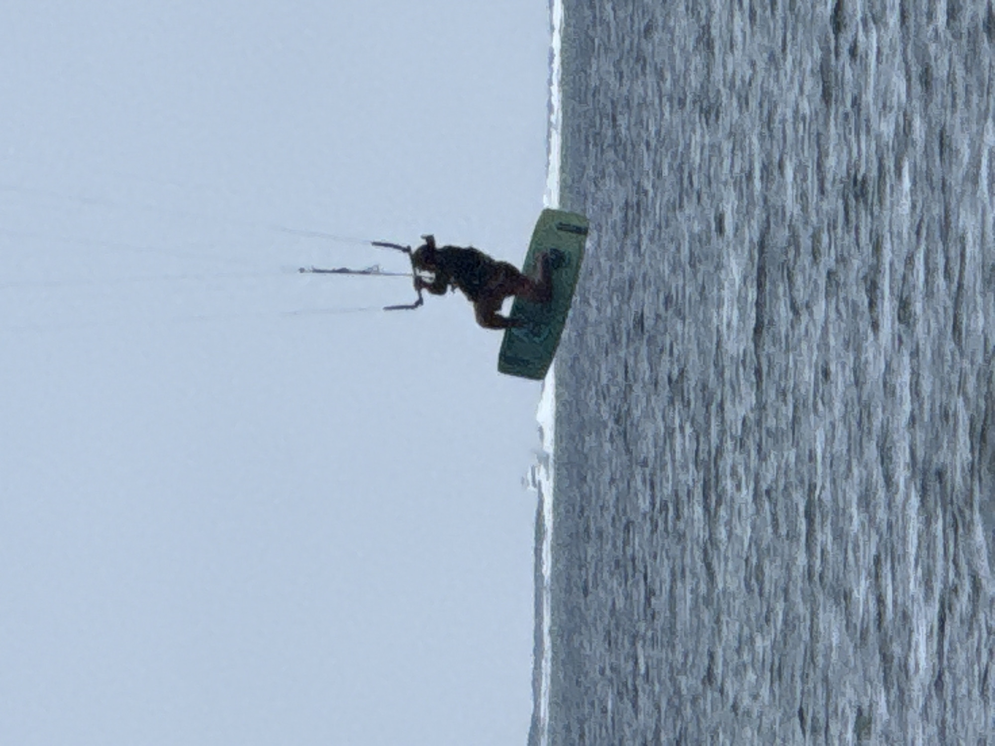 A kitesurfer is captured mid-air, suspended above the choppy gray water of the ocean. The kitesurfer, silhouetted against a light blue sky, is positioned on a green surfboard, holding onto a control bar connected to a kite by thin lines. The kitesurfer appears to be wearing dark clothing. In the background, a white boat navigates through the water, creating a white wake. The horizon line is marked by a low, indistinct landmass. The scene is set in the city of Denpasar, Indonesia, suggesting a coastal location known for watersports.