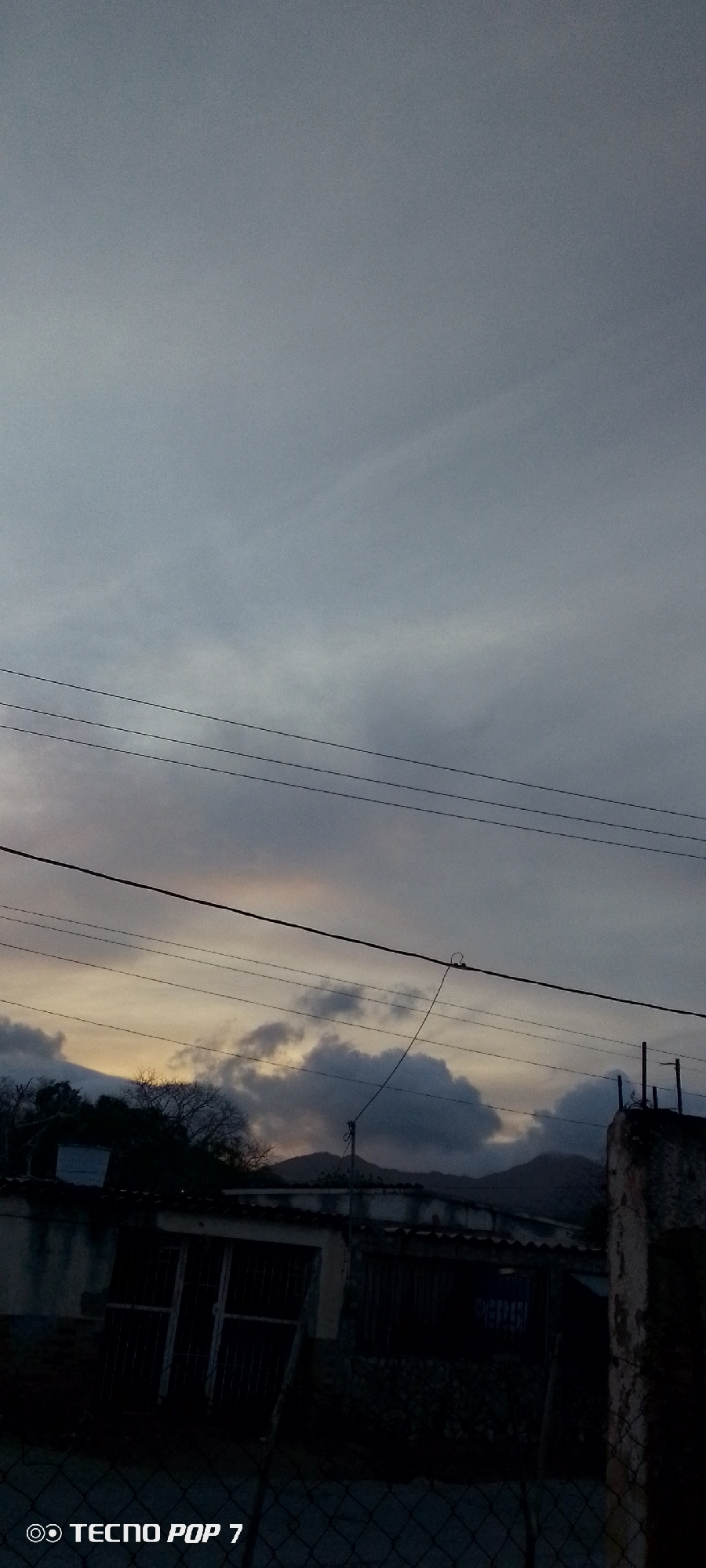The image depicts a scene in Altagracia, Venezuela. The foreground shows a section of a chain-link fence. Behind the fence are buildings, with dark shadows obscuring many details. The sky is overcast, with a few patches of brighter light peeking through the clouds. Power lines stretch across the sky above the buildings. In the background, there are hills or mountains. The image was taken with a Tecno Pop 7 phone.