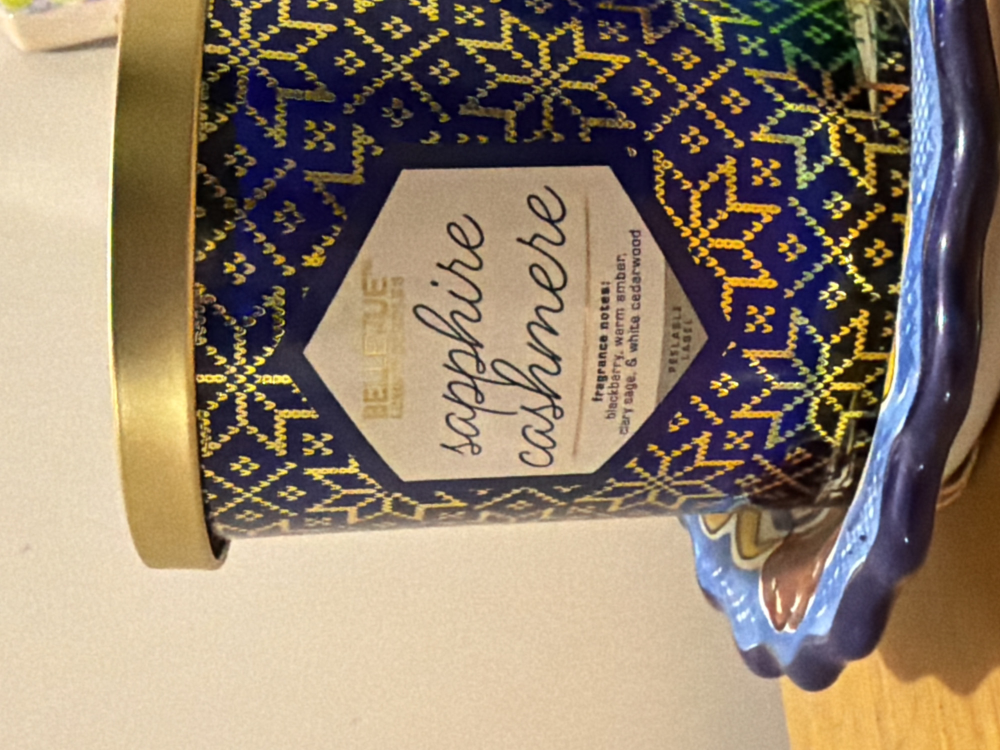 A close-up shot features a blue cylindrical container with a gold rim, resting on a blue ceramic dish. The container is patterned with small, repeating gold geometric designs on a dark blue background. A central beige hexagonal label reads "BELLEVUE" above "sapphire cashmere" in cursive, with fragrance notes listed below: "blackberry, warm amber, clary sage, & white cedarwood". The label also includes the text "PEELABLE LABEL". The dish is partially visible, with a decorative blue border. The background is a beige wall, suggesting an indoor setting. The location in the city of Santa Clara Ocoyucan, Mexico, is not discernable from the provided image.