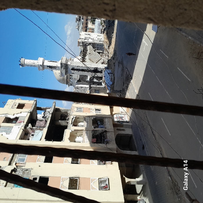 This image depicts a scene of extensive urban destruction in the city of Khan Yunis, Palestinian Territory, viewed from an elevated vantage point through vertical metal bars in the immediate foreground. To the left, a multi-story building, light-colored and of residential appearance, exhibits severe structural damage, particularly across its upper and middle floors, with large sections collapsed, interiors exposed, and numerous windows shattered. Fabric items are suspended from several damaged balconies. In the background to the right, a mosque, identifiable by its minaret and dome, has suffered significant structural collapse, reducing its main edifice to rubble while the minaret remains partially upright but damaged. A paved, two-lane road with faded white lane markings traverses the mid-ground. A lone individual, dressed in dark clothing, walks along the center of this road from left to right. The sky is clear and blue. Overhead utility wires crisscross the upper portion of the frame. Debris is scattered around the damaged structures, though the visible road surface is largely clear.