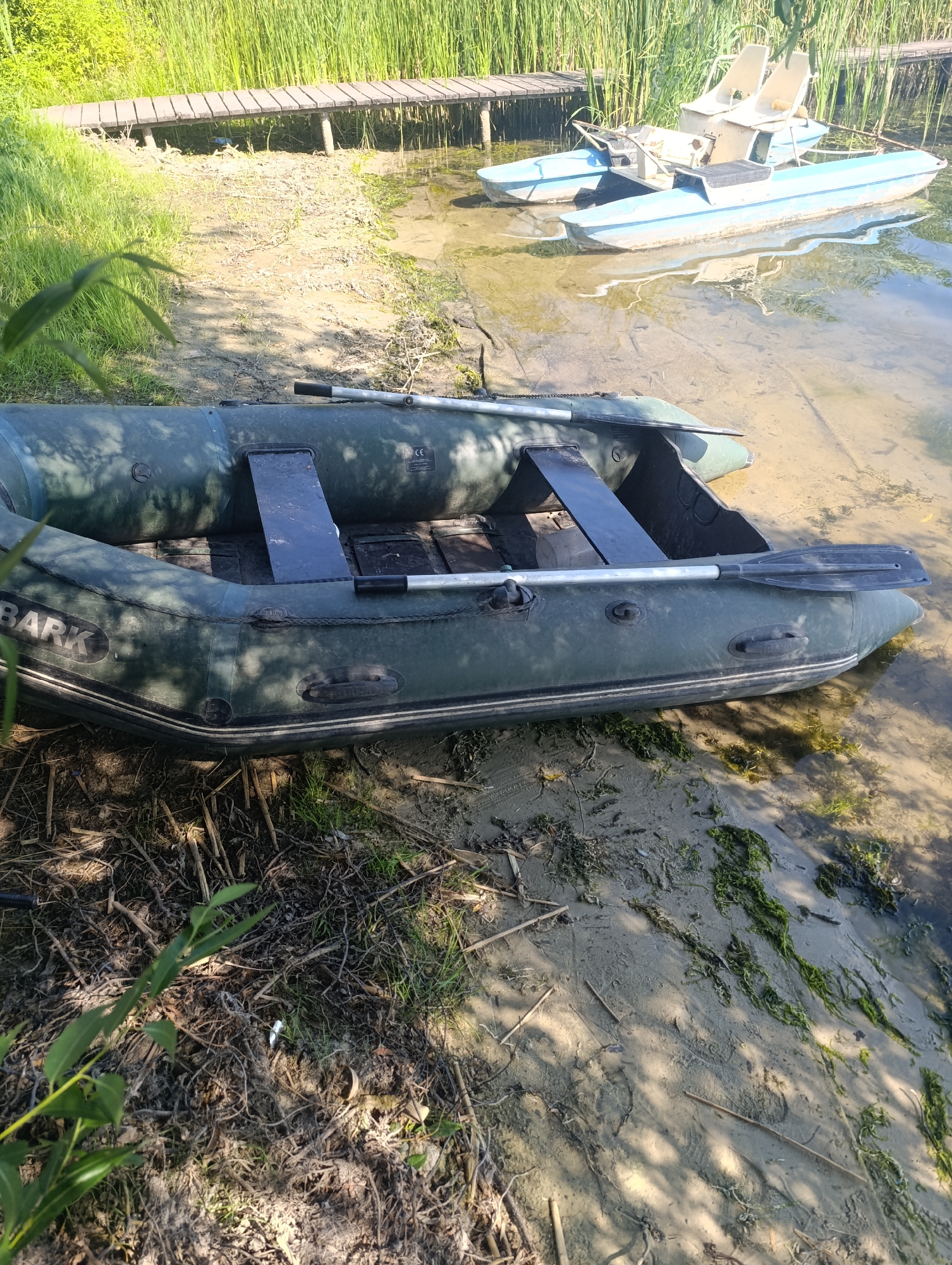 An inflatable raft, dark green in color, rests on a sandy shore at the edge of a body of water. Two oars are positioned across the raft. In the background, a wooden boardwalk extends over the water, and tall reeds line the shore. A blue paddle boat with two seats is visible on the water near the boardwalk. The environment is natural, with sunlight filtering through trees, casting shadows on the ground. The scene appears to be located in Bulatselivka, Ukraine, a rural area with a focus on outdoor recreation.