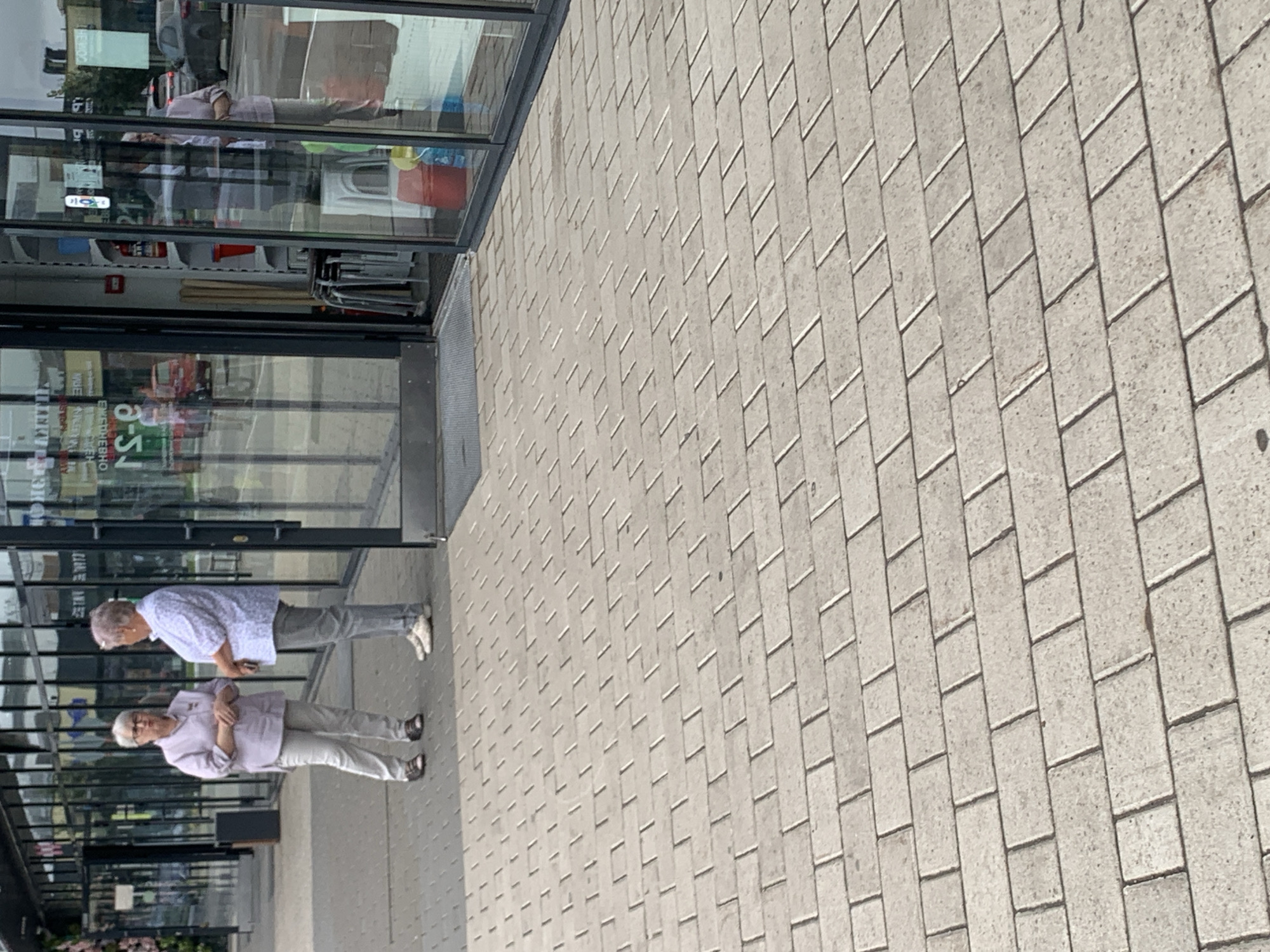 Two elderly women are standing on a paved walkway in front of a store with large glass windows. The woman on the left is wearing a light purple shirt and light gray pants. She has short gray hair and glasses, and is looking at the woman on the right. The woman on the right is wearing a white patterned shirt and gray pants. She has short gray hair and is looking at the woman on the left. The store appears to be a retail establishment. The walkway is made of light-colored rectangular paving stones. The background shows reflections in the store windows, including cars and other buildings. The scene suggests a casual encounter or conversation outdoors.