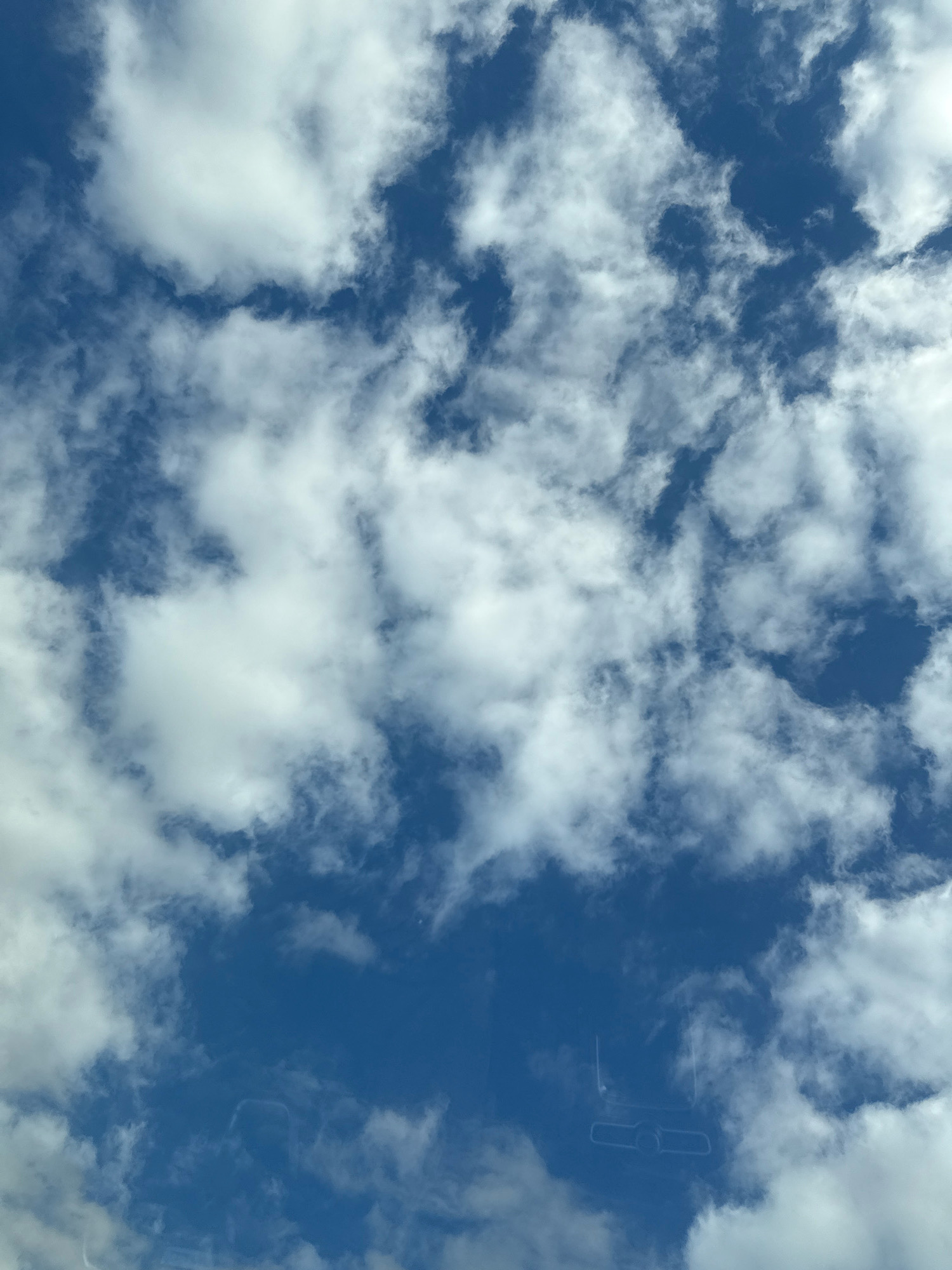 The image is a vertical, full-frame shot of a blue sky filled with scattered white clouds. The clouds vary in density, with some appearing wispy and others thicker and more opaque. The sky is a deep blue, indicating daytime. In the lower portion of the image, there is a faint reflection on what appears to be a windshield, showing the outline of a rearview mirror and possibly other interior elements of a vehicle. There are no people or distinct objects visible besides the clouds and the reflection. The scene is an outdoor environment, likely captured from inside a car looking upwards. The location in Herzberg, Germany is not discernible from the visual information provided in the image.