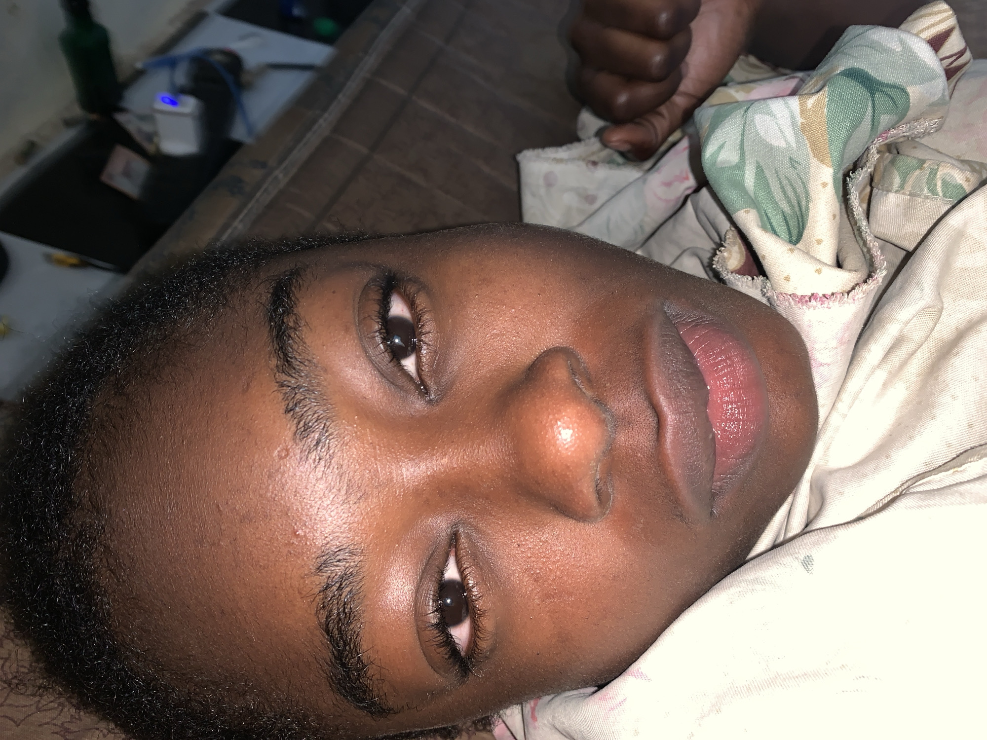 A close-up, tilted view shows a person with dark skin lying down. The person's eyes are partially open. The face occupies most of the frame. The person lies on bedding with floral patterns and a patterned brown surface beneath. In the background, there is a table or shelf with various items, including a green bottle, a white object with a blue light, and other indistinct objects. The location is Rafin Par, Nigeria. The scene depicts a person lying down with personal belongings nearby.