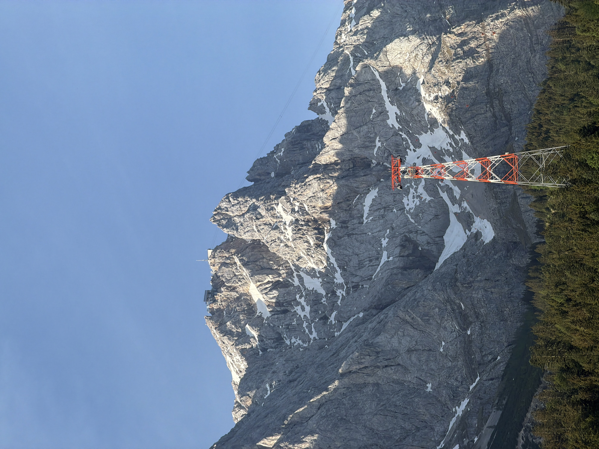 A low-angle, upward-facing shot depicts the Zugspitze mountain in Grainau, Germany. The mountain is primarily gray rock, with patches of snow clinging to its surface. An orange and white lattice tower is perched on the mountainside. Dark green trees grow to the right of the tower and on the right edge of the frame. The upper left portion of the image is a clear, light-blue sky. A small structure is visible near the peak of the mountain. A cable line runs from the small structure to the top of the mountain.