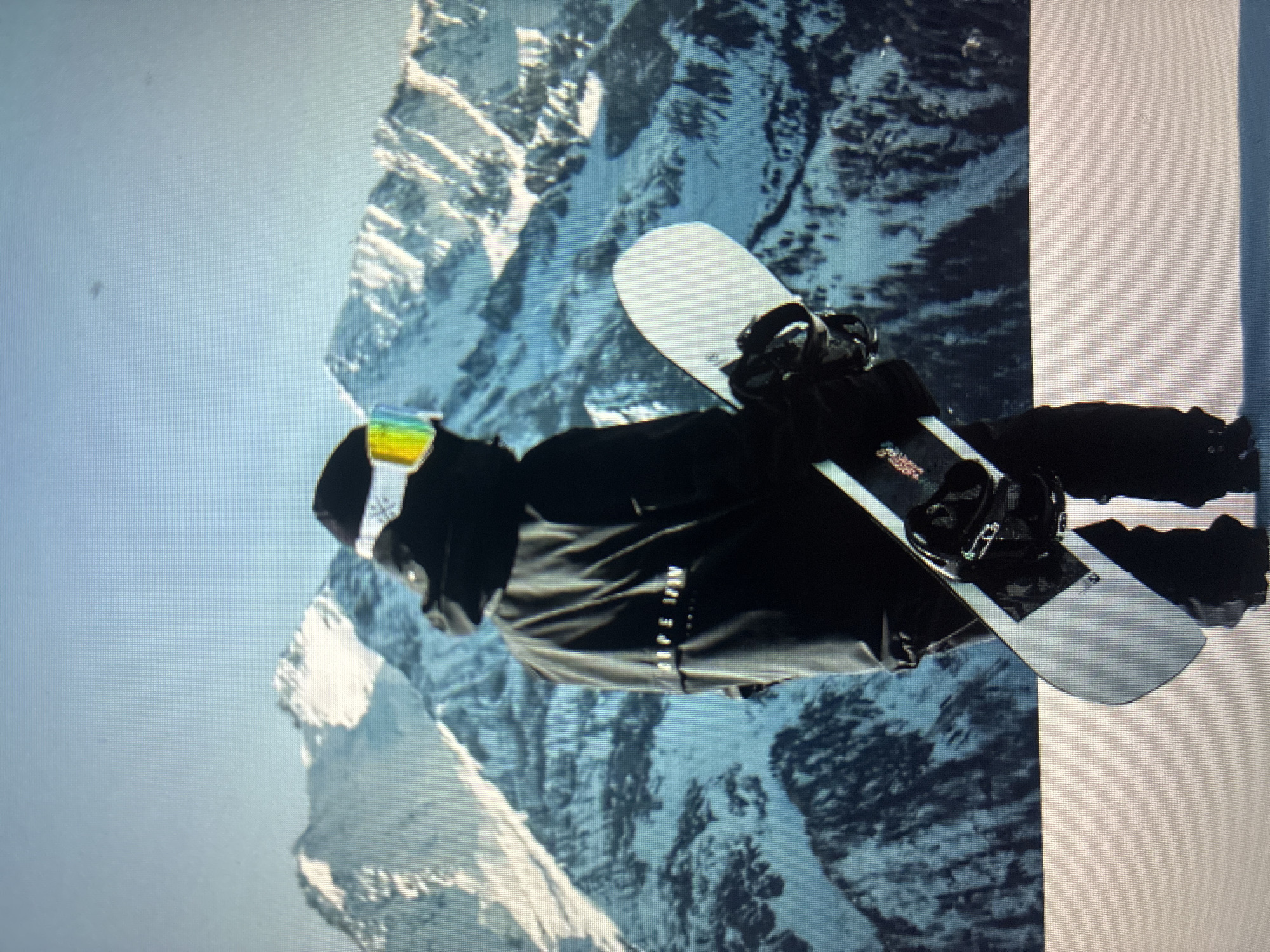 The provided image, rotated 90 degrees clockwise, depicts a snowboarder standing upright in a majestic, snow-covered mountainous landscape, possibly in Asenovgrad, Bulgaria. The central figure is a person dressed in dark winter attire, including a jacket, pants, and gloves. Their face is obscured by a black balaclava or neck gaiter, and they wear goggles with a vibrant, rainbow-tinted lens. The snowboarder is holding a white snowboard horizontally across their body, showcasing its black bindings.
The background is dominated by rugged, snow-capped mountains with dark, rocky outcroppings, indicating a high-altitude setting. The sky above is clear and bright, suggesting a sunny day. On the right side of the frame, a stark white, expansive snowfield or structure is visible, where the snowboarder appears to be standing.
Notable details include the bright lighting, crisp mountain air implied by the clear visibility, and the dynamic pose of the snowboarder. Faint, light-colored text is visible on the snowboarder's jacket, but not clearly legible. The image displays a grid pattern and pixelation, characteristic of a photograph taken of a digital screen rather than a direct digital file.