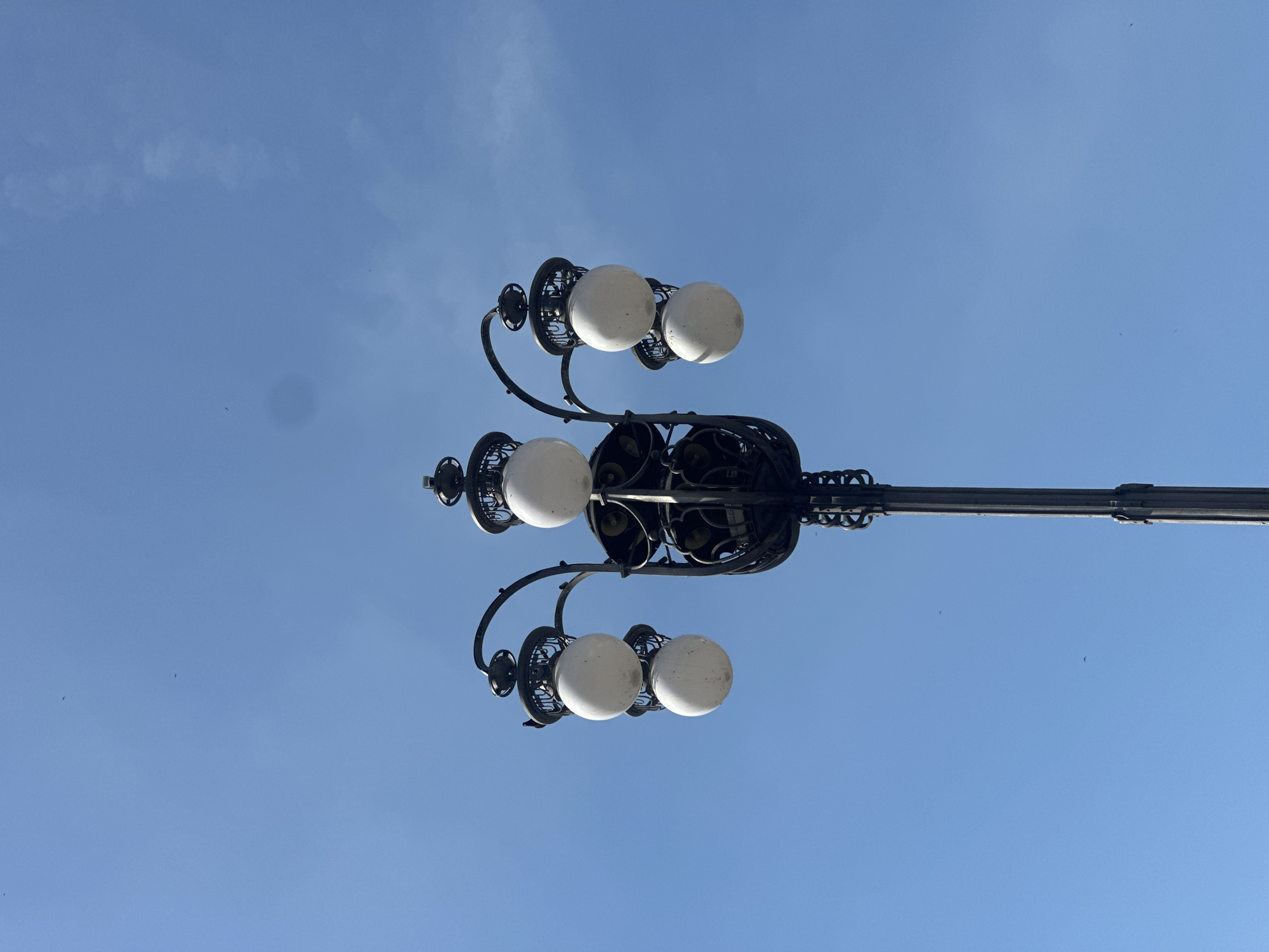 An ornate, dark-colored streetlamp with multiple globe-shaped lights stands prominently against a clear blue sky. The lamp has a central pole and several curving arms, each holding a white, frosted globe light. The globe lights are enclosed within decorative metal cages. The streetlamp is the primary subject, and the background is an expansive, cloudless sky. There are a few small dark specks visible in the sky, possibly birds. The image is taken from a low angle, looking upwards at the streetlamp.