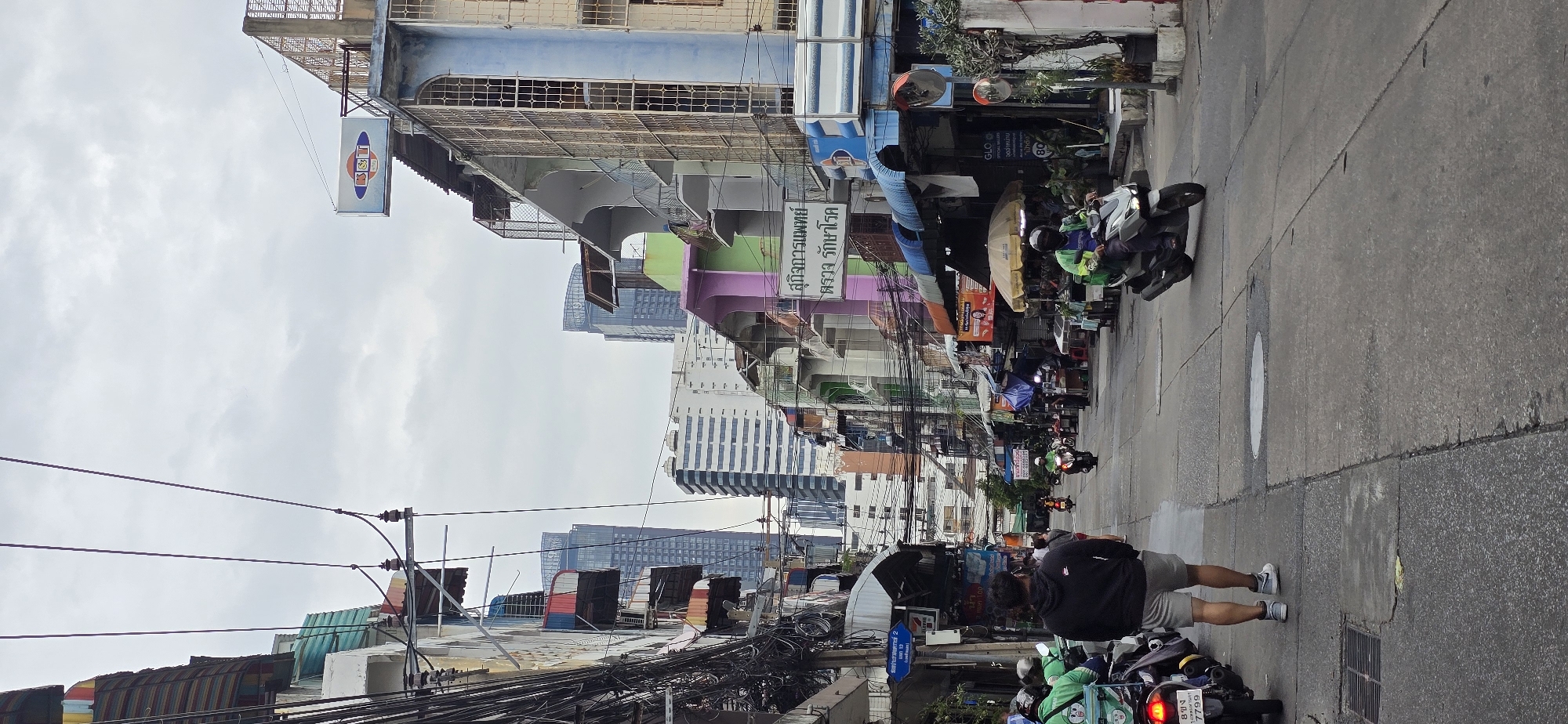 The image captures a bustling street scene in Bangkok, Thailand, likely a market or commercial district, characterized by narrow buildings, dense overhead electrical wires, and a mix of traditional and modern architecture. A person in a black hoodie and grey shorts walks away from the camera down the center of the paved street. On the left, several motorcycles, some with green-clad riders and delivery boxes, are parked. On the right, a motorcycle with a rider wearing a green jacket and helmet is in motion. Shops and stalls line both sides of the street, with awnings providing shade. Signs in Thai are visible on the buildings, including one that appears to be for a medical clinic. In the background, tall modern skyscrapers rise above the lower structures, indicating a dense urban environment. The sky is overcast, suggesting a cloudy day.