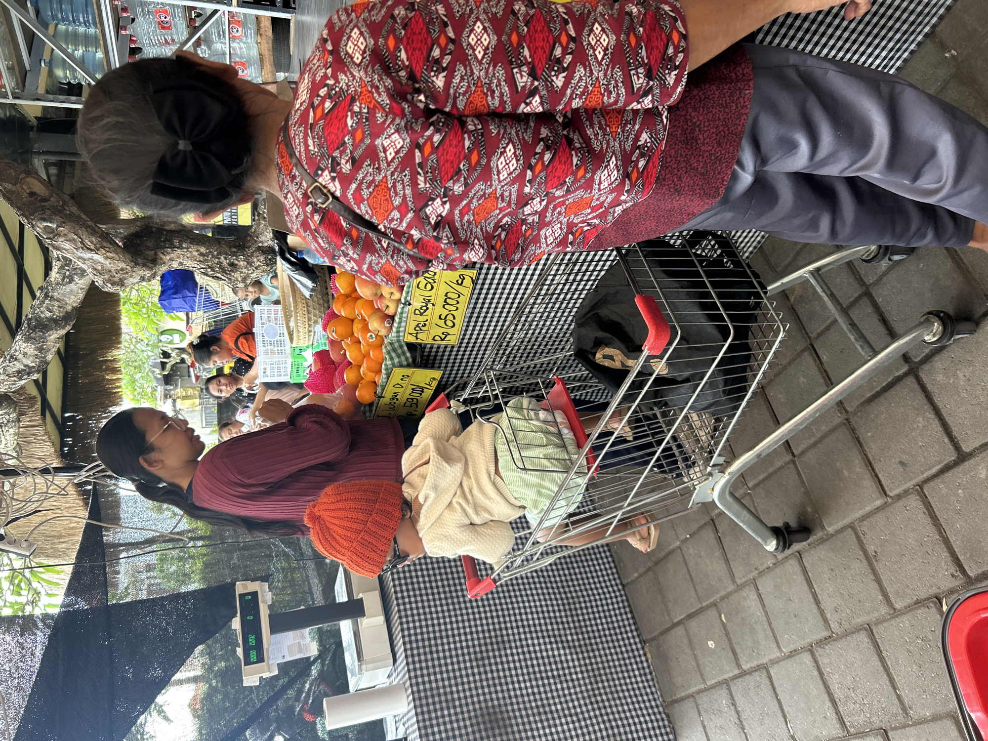In Sukawati, Indonesia, at a market stall, a woman with glasses and a maroon sweater stands at a scale. To her left, a toddler wearing an orange knitted hat, sunglasses, a cream-colored hoodie, and striped shorts sits in a shopping cart. In front of the woman are fruits, including oranges and apples, with a price sign reading "Apel Royal Gala Rp 65.000/kg". Another woman, seen from the back, wears a red patterned shirt and dark pants. The market environment is visible with various stalls and shoppers in the background. The ground is paved with gray bricks.