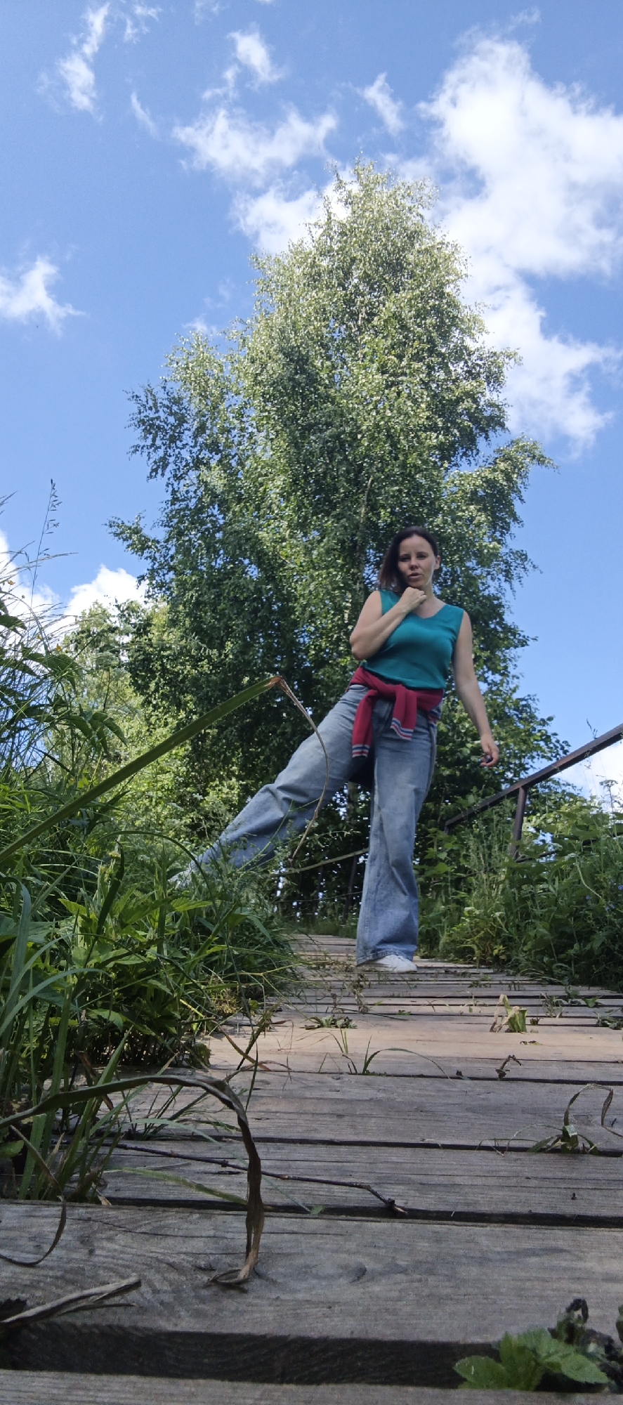 A low-angle full shot captures a woman on a wooden boardwalk surrounded by lush greenery under a bright blue sky with scattered white clouds. The woman, who appears to be of East Asian descent and has brown hair, is wearing a turquoise sleeveless top, baggy blue jeans, and white sneakers. A red sweater with white stripes is tied around her waist. She is standing with her right leg extended forward, her body angled slightly towards the left, and her left hand touching her chin. The boardwalk is weathered and appears to be in a natural setting, with grass and small plants growing between the wooden planks. A metal railing runs along the right side of the boardwalk. The background features a large deciduous tree, its branches reaching towards the sky. The image is taken outdoors in what appears to be a park or natural area.