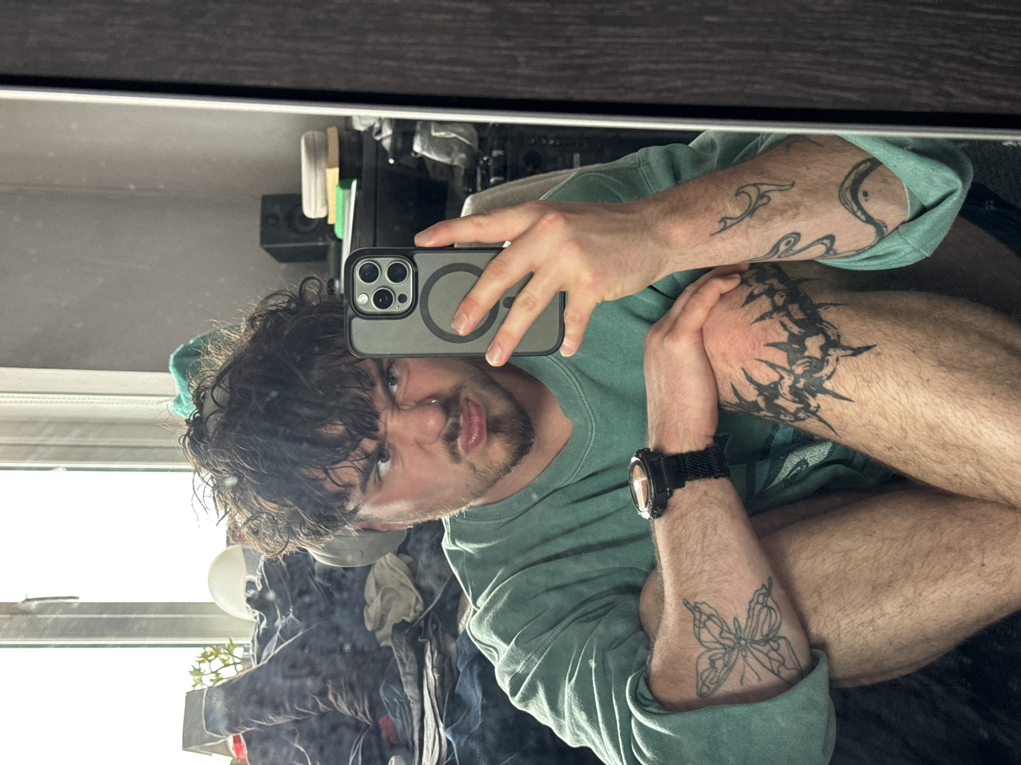 A person with curly dark hair, a mustache, and a nose ring is taking a selfie in a mirror with a gray phone. The person is wearing a green sweatshirt. Tattoos are visible on the person's arms and legs, including a butterfly on the forearm and a barbed wire design on the leg. A black watch is on the person's left wrist. The background includes a window with daylight coming through, a pile of clothes, a white lamp, and some plants on a windowsill. The location is Unknown, Unknown.