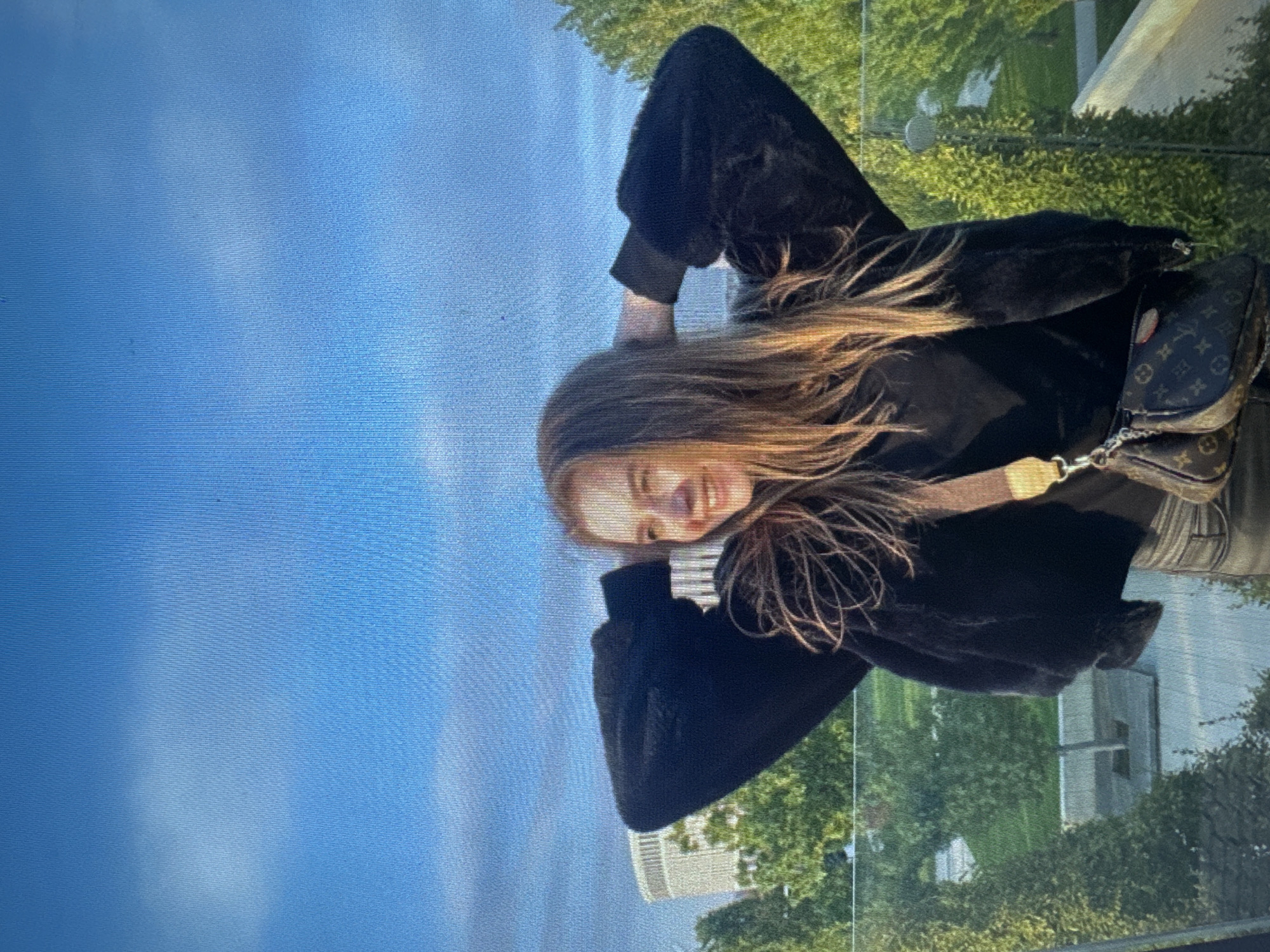 A young Caucasian woman stands outdoors with her hands behind her head, smiling and looking to her left. She is wearing a black faux fur bomber jacket over a black top and dark pants. A small designer crossbody bag with a brown and black monogram pattern hangs from her shoulder. Behind her, a blurred background shows a modern white building with a curved facade, lush green trees, and a clear blue sky. The woman is positioned on a raised platform or balcony, with a glass railing in the foreground. The lighting suggests it is daytime, possibly late afternoon due to the warm tones in the sky.
