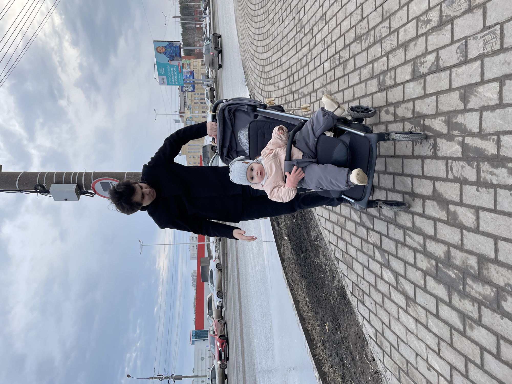 A person in a black coat and dark pants stands on a brick-paved sidewalk next to a black stroller occupied by a baby wearing a gray hat and light pink jacket. The stroller faces right in the frame. A narrow strip of dirt or mulch borders the sidewalk. To the left, cars are parked along a street, and a billboard is visible in the background. Based on the billboard and surrounding cityscape, the location appears to be in Izhevsk, Russia. The sky is overcast.
