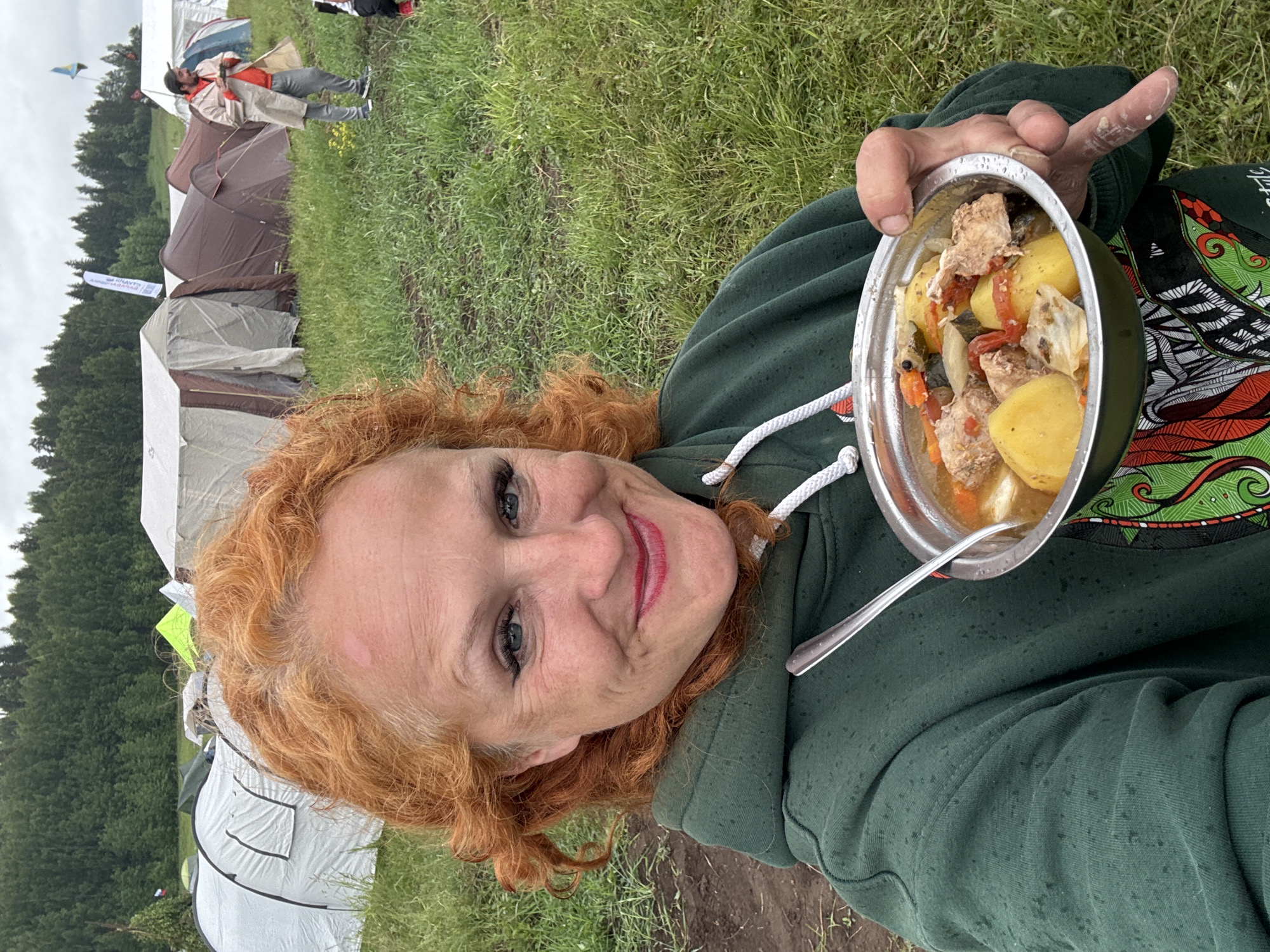 A woman with vibrant red, curly hair and blue eyes smiles warmly at the camera in a selfie taken outdoors. She is wearing a dark green hoodie with a white drawstring and a colorful, abstract graphic design on the right shoulder and sleeve. She holds a metal bowl filled with a hearty stew, containing large pieces of potato, meat (likely chicken or pork), and chopped vegetables such as tomatoes and carrots. A spoon rests in the bowl.
The setting appears to be a camping or outdoor festival in Большая Кивара, Russia. In the background, several tents of various colors (grey, brown, white) are pitched on a grassy field. A line of dense green trees forms a forest in the distance under an overcast sky, indicating it is daytime. Another person, dressed in a light-colored trench coat and dark pants, is visible standing among the tents further back. The ground is green with grass, and the woman's hoodie shows small water droplets, suggesting damp conditions or recent rain.
A white flag or banner with partially visible Cyrillic text, possibly "НПАУТТ" and "БАСКАНДИ," stands upright in the middle-ground. The woman's joyful expression suggests she is enjoying her meal and the outdoor experience.
