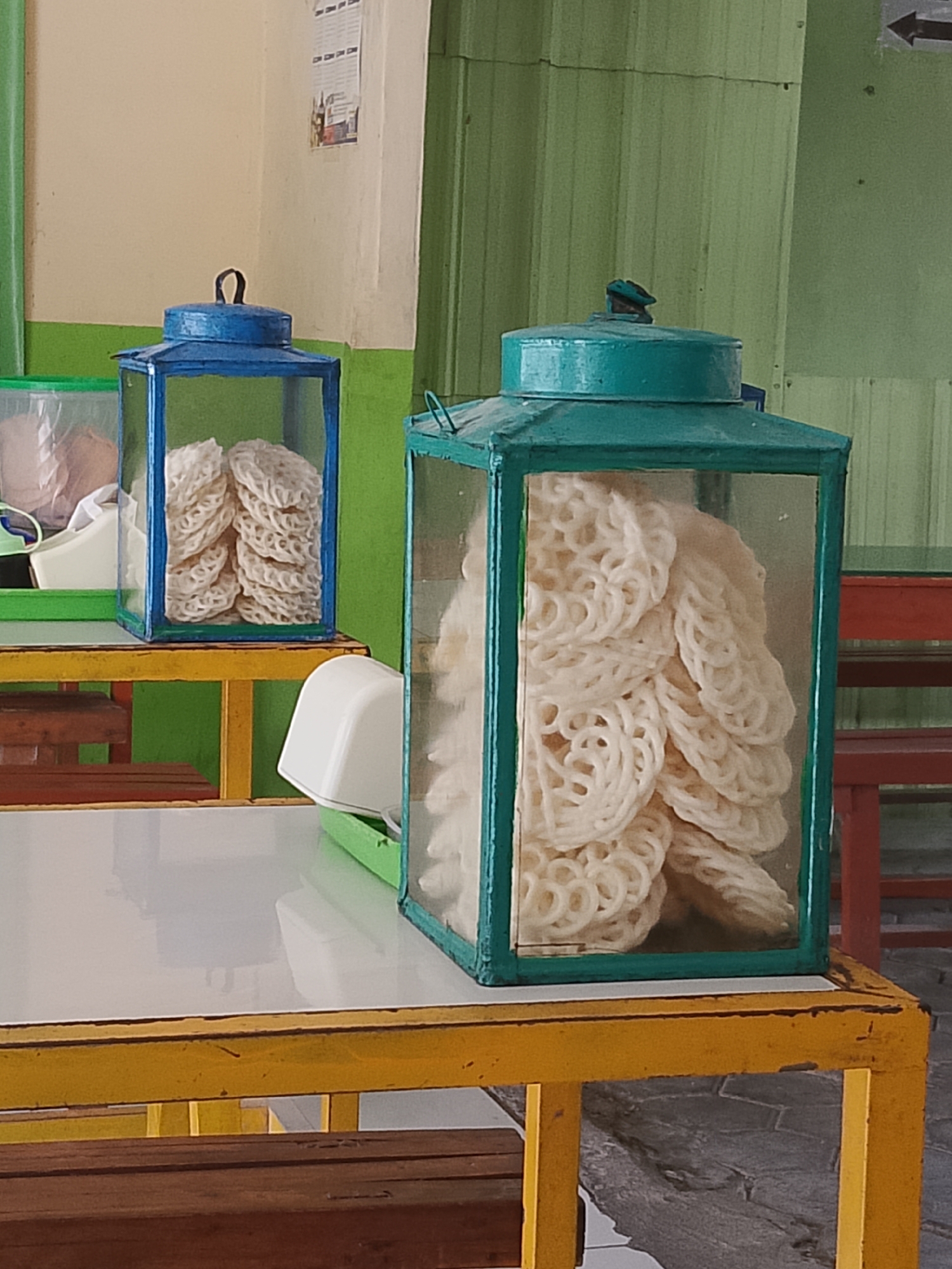 The image shows two decorative lanterns filled with white, circular crackers. The lantern on the left is blue, and the lantern on the right is teal. Both lanterns are made of glass and metal and are sitting on yellow tables. The tables are in front of a green wall and a beige wall, which appears to be indoors or in a covered outdoor area. The cracker display is likely in a food stall or restaurant. Based on the context of Balun, Indonesia, this scene depicts a street food vendor displaying traditional crackers for sale.