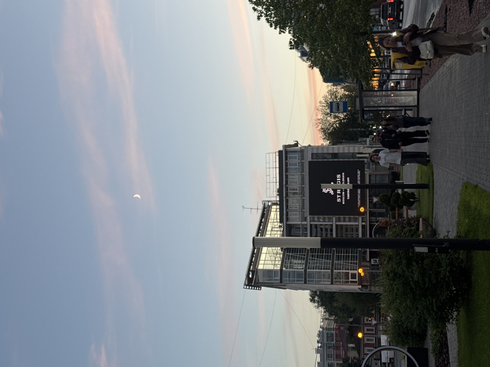 A wide-angle, eye-level outdoor shot depicts a street scene in Nizhny Novgorod, Russia during dusk. The sky is a pale blue with faint pink streaks from the setting sun, and a sliver of the moon is visible. In the mid-ground, a modern glass-fronted building with a prominent digital billboard displays the text "STRATIGIS" and what appears to be a logo above the words "MOSCOW - OLSKAYA" and a web address. A bus stop shelter with a blue bus sign is located to the right of the building. A sidewalk runs in front of the building, with four individuals walking towards the camera. The person on the far right is a woman carrying a yellow insulated bag on her back and a white purse on her shoulder, while looking at her phone. To her left, a group of three individuals, two men and one woman, are walking together. Further left, a large green bush is visible in the foreground. Streetlights and trees line the sidewalk. A street with moving vehicles is to the right of the bus stop. The overall atmosphere is calm and urban.