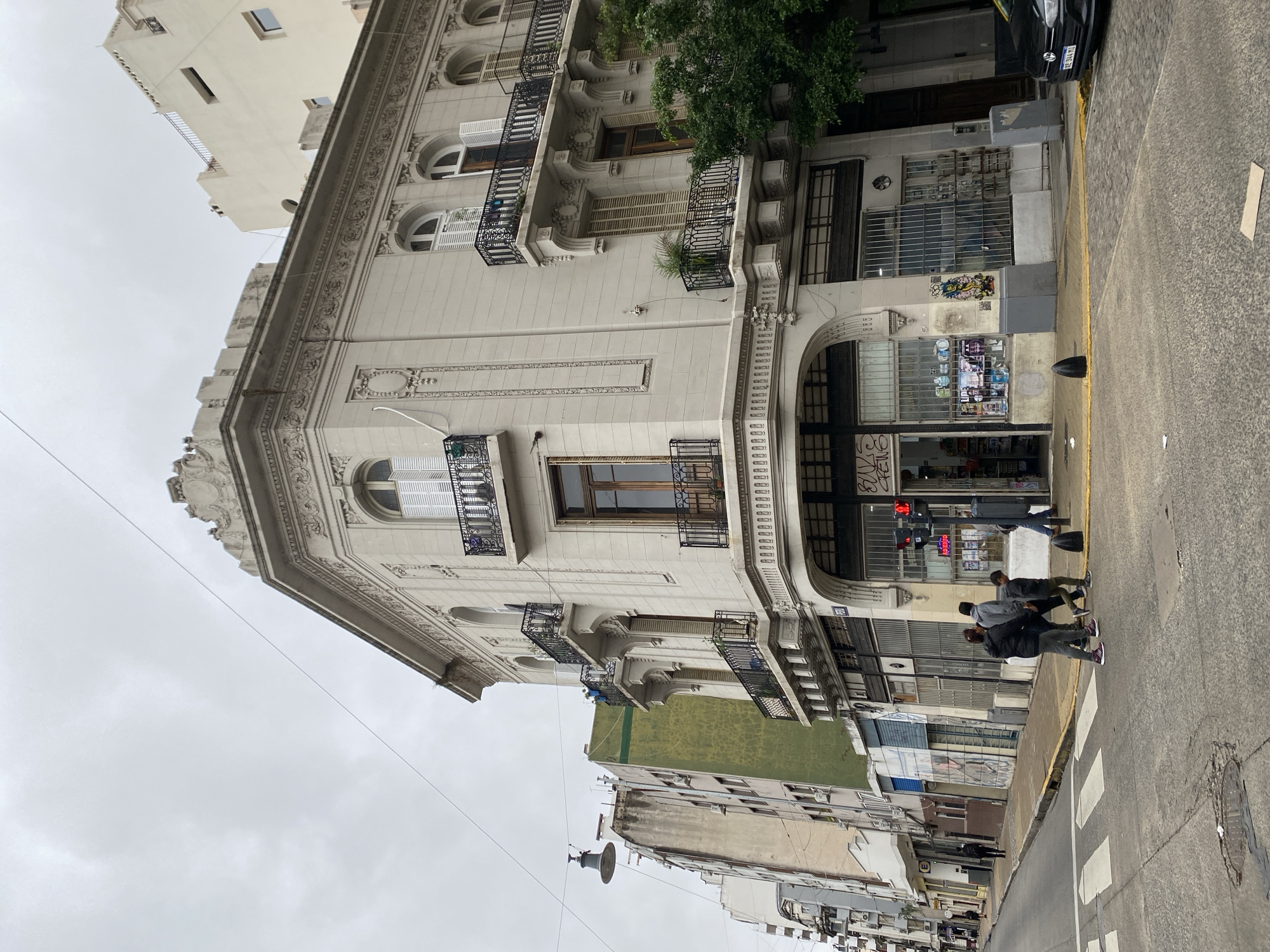 The photograph presents a street corner in Buenos Aires, Argentina, under an overcast sky. The focal point is a multi-story building with ornate architectural details, including decorative moldings, balconies with wrought-iron railings, and arched windows. The building's facade is light in color.
A commercial establishment occupies the ground floor, identifiable by its display windows and security bars. There is signage that can be seen behind the window. A group of people is crossing the street in the foreground, using a pedestrian crossing. Their attire suggests casual, everyday wear.
The street itself appears to be asphalt and has a yellow curb. Additional buildings line the street in the background, showcasing varying architectural styles and conditions. An overhead cable stretches across the street, partially obscuring the sky. There is a parked car by the curb in the background.