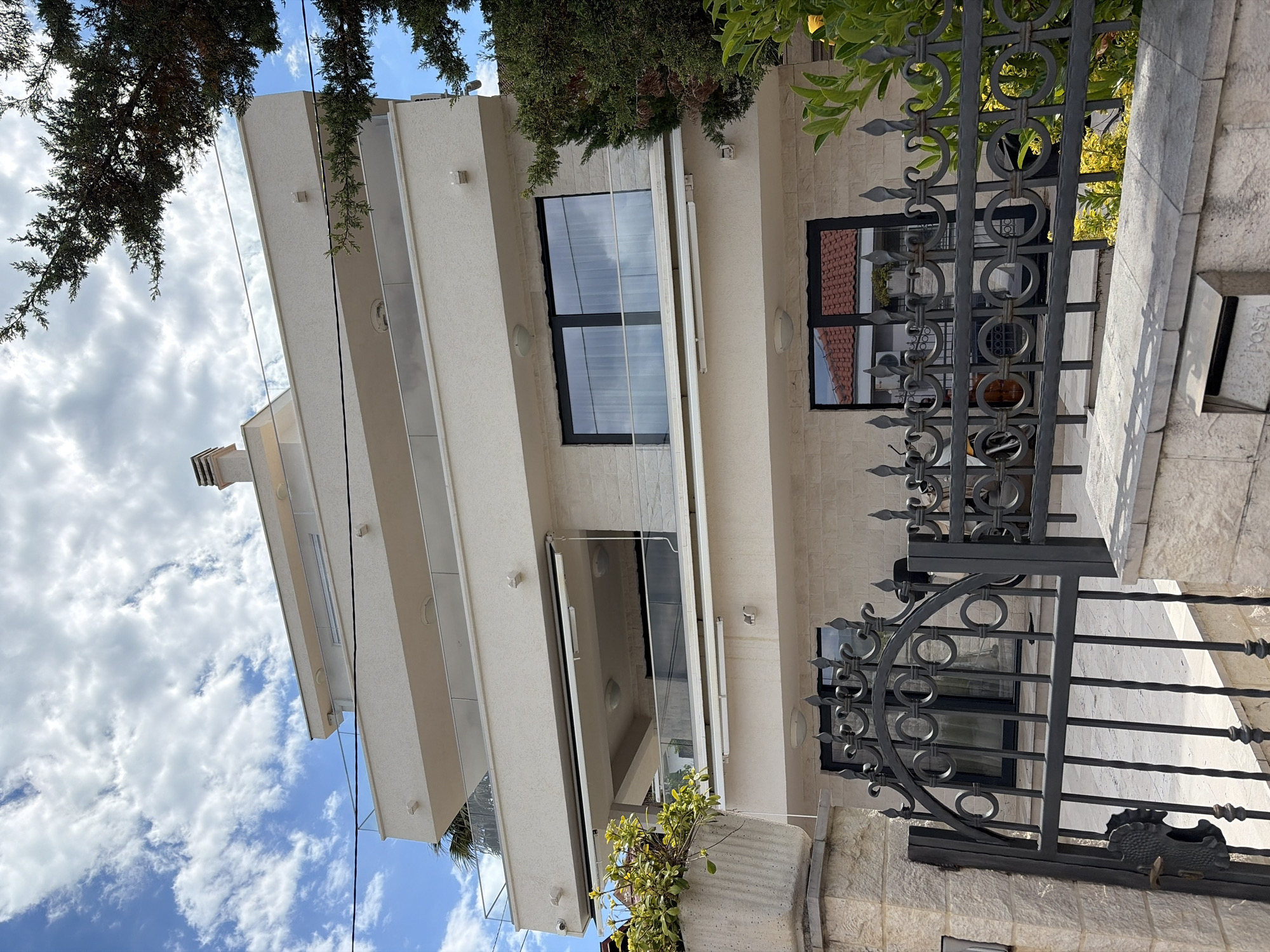 A modern building with staggered balconies and large windows is visible behind a dark metal fence. The building's facade is light-colored, possibly stone or concrete. Some windows reflect the sky, indicating they are closed. The fence has decorative circular and spear-shaped elements. Above the building, there's a partial view of trees and a blue sky with clouds. A mailbox with the word "POSTA" is near the fence on the right. The location appears to be Krašići, Montenegro, judging by the style of the building and fence. There are no people visible in the image.