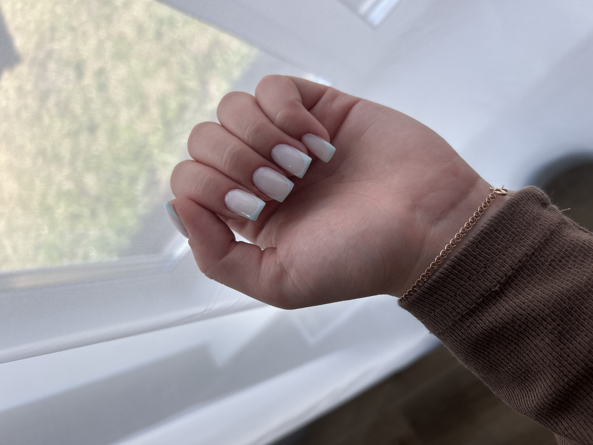 A close-up shot captures a person's left hand, gently curved and positioned centrally within the frame. The hand features light-toned skin, and its fingernails are manicured in a modern French style. Each nail has a translucent, milky-white base with distinct, squared-off tips painted in a soft light blue or mint green color. A delicate, gold-colored chain bracelet adorns the wrist, partially visible on the right side of the hand. The sleeve of a brown, ribbed, long-sleeved garment, likely a sweater or shirt, covers the forearm and wrist area.
The scene is set indoors, adjacent to a window. A sheer, white, subtly textured curtain or blind hangs, diffusing the bright daylight streaming in from outside. Beyond the curtain, an out-of-focus background reveals blurry green foliage, suggesting trees or bushes in a natural outdoor environment. The lighting indicates it is daytime. The image primarily highlights the hand, its manicure, and the bracelet, suggesting a presentation or detail shot. No specific activity is taking place beyond this display. There is no visible text in the media file. The location context for this image is Chaikovsky, Russia.
