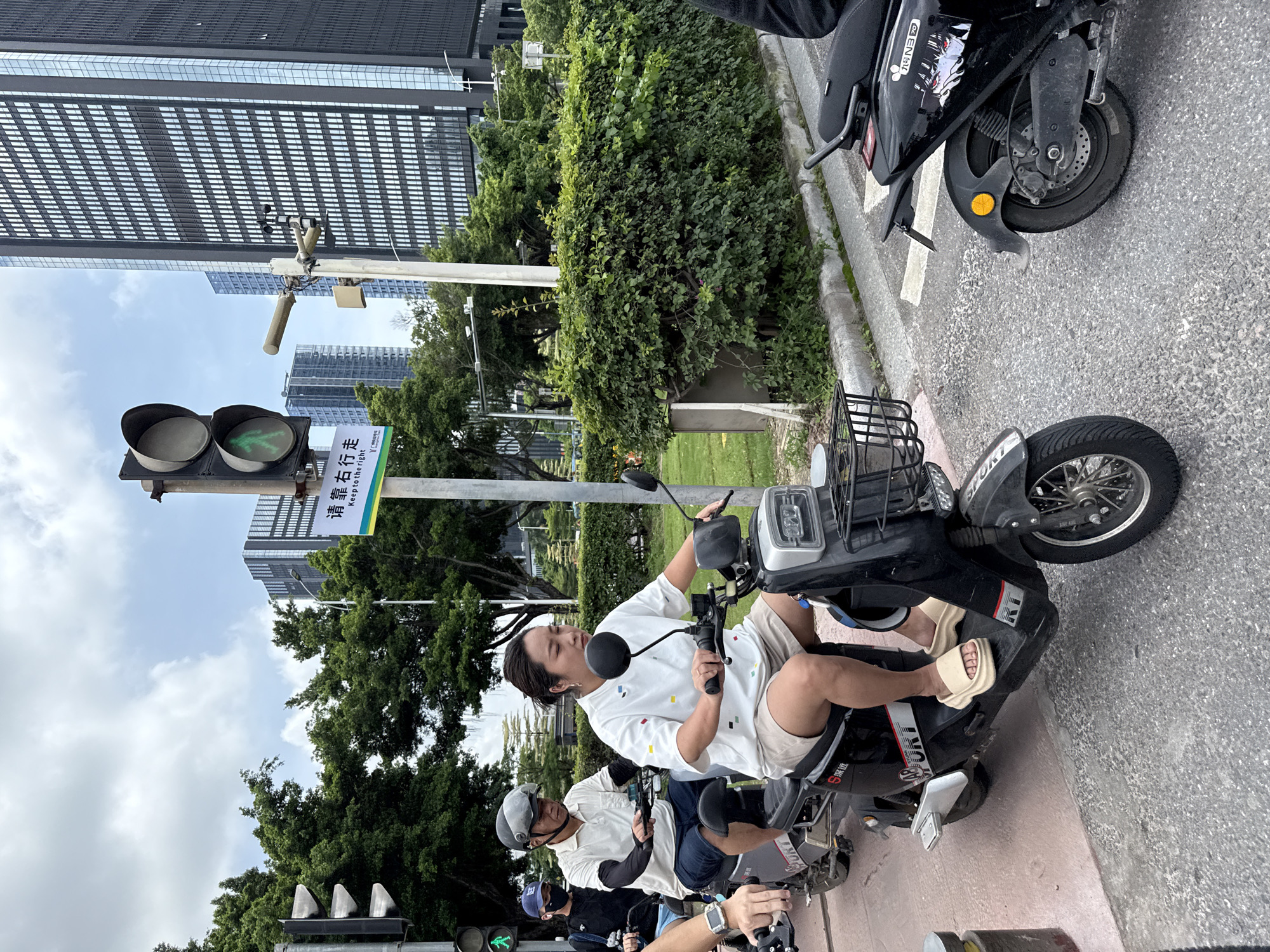 In Haizhu District, China, this image captures a street scene with several individuals on electric scooters at a traffic light. The foreground features a woman on a black scooter, wearing a white short-sleeved shirt with a geometric pattern and light brown shorts. She is looking forward and has her hands on the handlebars. Behind her, a man in a white polo shirt and blue shorts, wearing a helmet, is also on a scooter. Further back, another person on a scooter wears a blue cap and a black shirt. The scooters are parked in front of a pedestrian crossing. A traffic light overhead displays a green walking person symbol, indicating it is safe for pedestrians and cyclists to proceed. In the background, modern high-rise buildings tower over lush green trees and manicured hedges, signifying an urban environment with a mix of nature and contemporary architecture. The sky is partly cloudy.