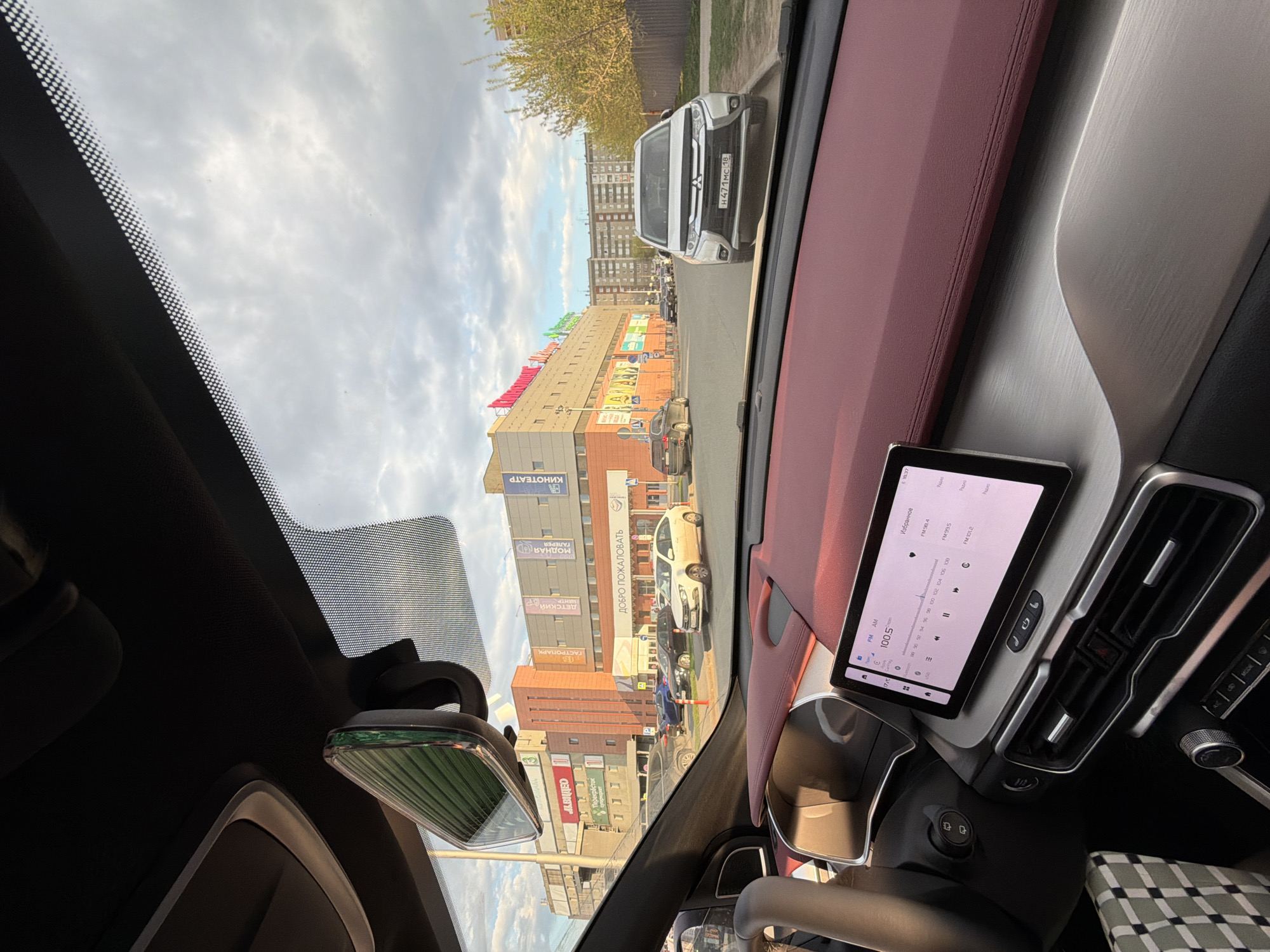 The image shows a view from inside a car looking out at a street in Izhevsk, Russia. The car's dashboard is visible, with a touchscreen display showing FM radio frequencies. Outside the car, there is a commercial building with several signs, including "Перекресток" (Perekrestok, a grocery store chain), "ГАСТРОПАРК" (Gastropark, a food hall), "Детский" (Detskiy, children's), "Модная Галерея" (Modnaya Galereya, a fashion gallery), "Добро Пожаловать" (Welcome), and "Кинотеатр" (Kinoteatr, cinema). Various cars are parked along the street, including a white Mitsubishi Outlander with the license plate "H471MC 18". The sky is cloudy.