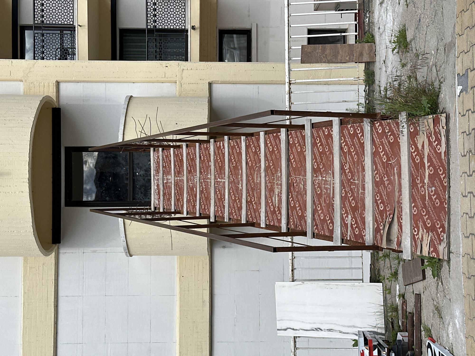 The image depicts the exterior of a building, likely in an urban setting, with ongoing construction work. Architectural details include windows with decorative latticework on the upper levels, and what appear to be arched or cylindrical structural elements. A scaffolding-like structure made of brown metal bars with plywood sheets (printed with the words "GTC FORMWORK") is visible in the foreground and seems to be used for construction. The ground level has white tiled walls. The background includes patches of overgrown plants. Construction materials like steel pipes, white marble tiles and other debris are also seen in the foreground. The location, city of Veliko Tarnovo, Bulgaria, is not deducible from the image.