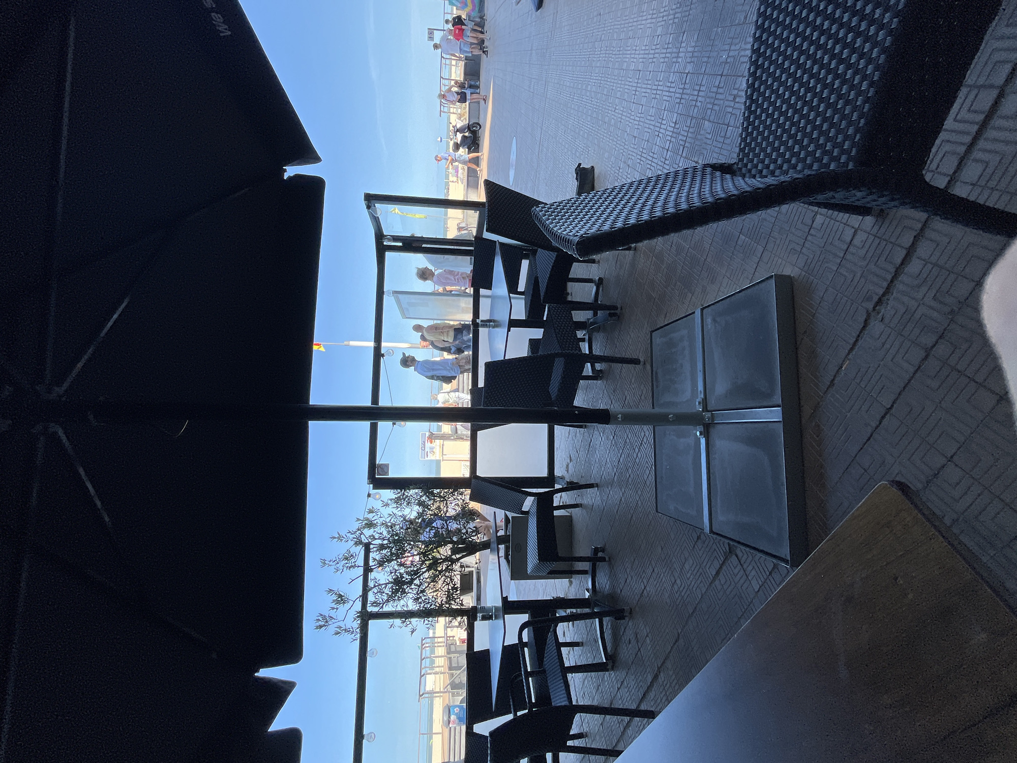 The image depicts an outdoor seating area at a beachside establishment in Ostend, Belgium. A large black umbrella partially covers the foreground, casting a shadow over several tables and chairs. Through the transparent partitions of the seating area, a view of the beach and promenade is visible. Several people are seen walking on the promenade and beach, and a scooter is parked nearby. The ground is paved with a textured tile pattern. The overall atmosphere suggests a casual dining or relaxation spot by the sea.
