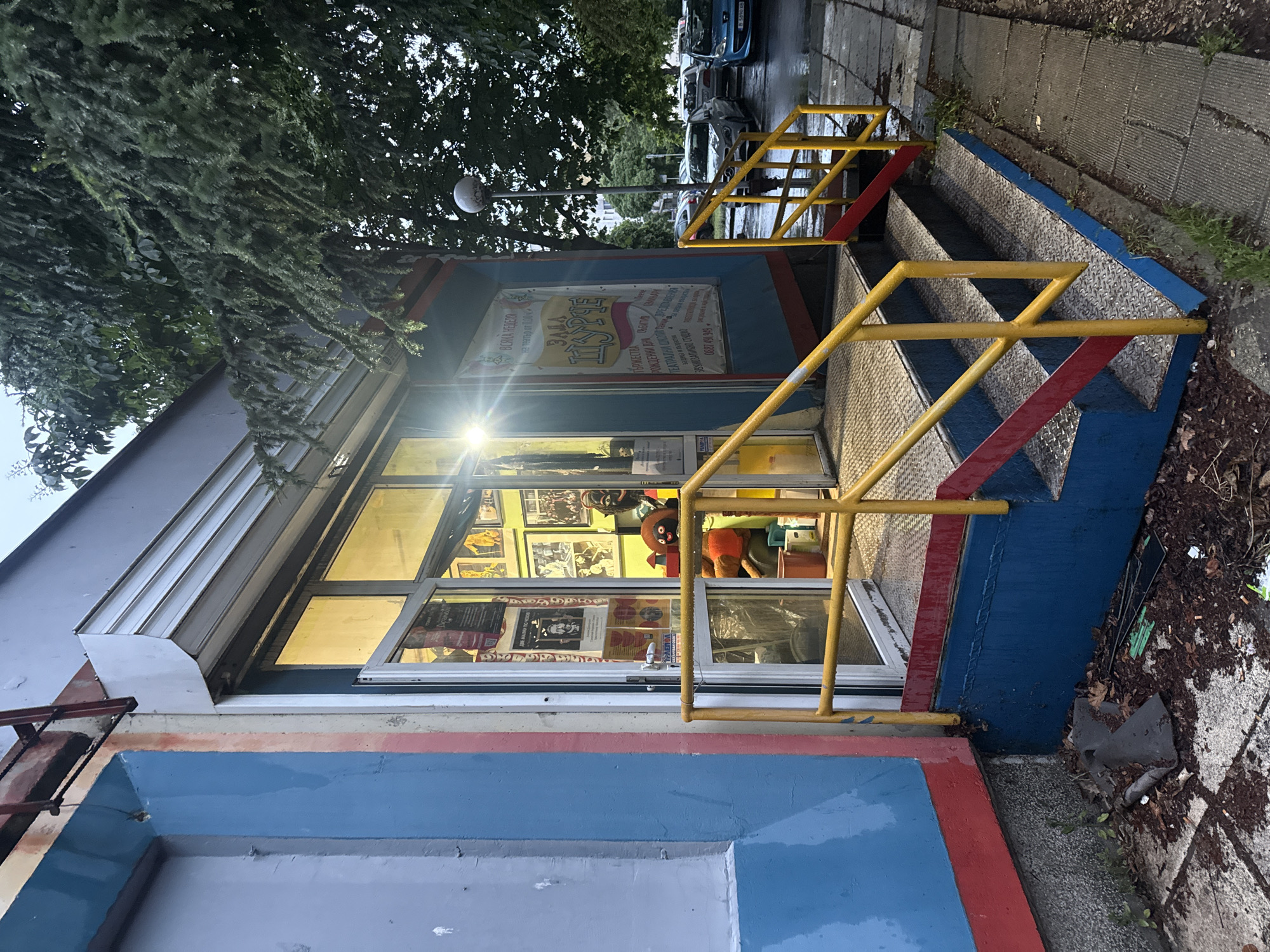 This medium-angle photograph captures a brightly lit building at what appears to be dusk or night, indicated by the strong glow from inside and the darker, muted tones of the sky and surroundings. The ground, including the sidewalk and steps, appears wet, suggesting recent rain.
The focal point is a small, rectangular building with a distinctive exterior painted in bands of blue, red, and gray. A prominent entrance features a set of steps leading up to a glass door. These steps are made of diamond-plate metal, bordered by blue and red accents, and equipped with bright yellow metal railings.
Through the large windows of the building, a brightly lit interior is visible, showing framed pictures on the walls and what appears to be a large, dark-skinned doll with a red hat on a shelf. Above the entrance, a large white sign with text in Cyrillic is displayed.
To the left of the building, dark green foliage from an overhead tree partially obscures the view. To the right, beyond the steps, a paved sidewalk leads to a street where several cars are parked, and a streetlamp is visible in the distance.
Visible text on the sign, in Bulgarian, lists services and information: "ЗАЛА ТЪРЖЕСТВА" (HALL CELEBRATIONS), "РОЖДЕННИ ДНИ СВАТБИ" (BIRTHDAYS WEDDINGS), "ТЕАТРАЛНИ ШКОЛИ" (THEATER SCHOOLS), "ЗВУКОЗАПИСНО СТУДИО" (RECORDING STUDIO), and "ПРЕДСТАВЛЕНИЯ" (PERFORMANCES). A phone number "0887 491 949" is also present. Another smaller text states "ДВЕ МАЛКИ ПО ЧЕХОВ на театър от 1.00", which translates to "TWO SMALL ONES BY CHEKHOV at the theater from 1.00", likely advertising a play or performance. The location context is Varna, Bulgaria.