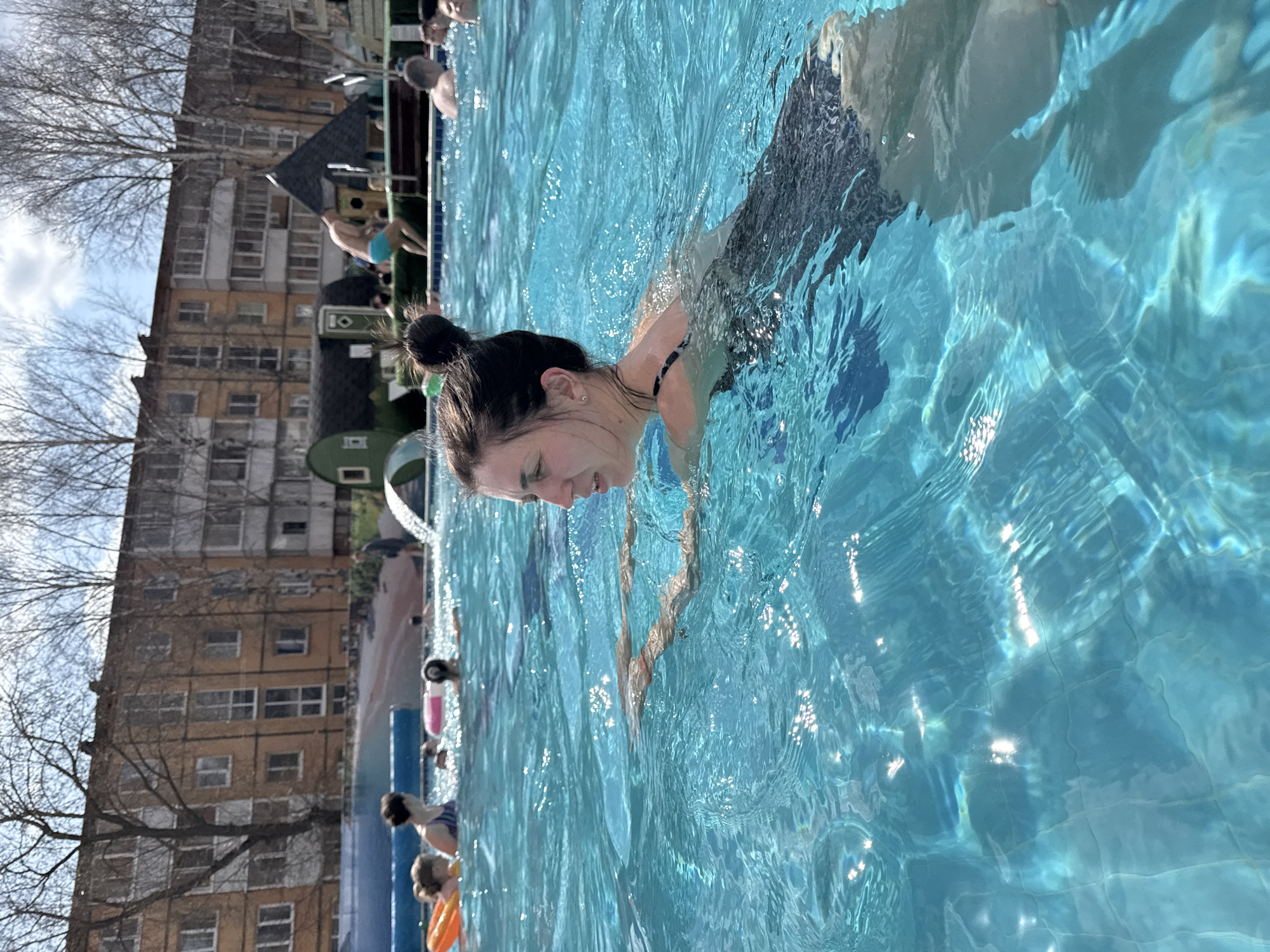 A medium shot depicts a light-skinned woman with dark hair pulled into a bun, swimming in an outdoor pool. She is seen from the chest up, with her face mostly submerged in the water, facing downward. The pool water is clear and reflects the sunlight.
In the background, other people are visible in the pool, and there is a building complex on the left side of the frame. The building appears to be a multi-story structure with yellow facades. The location is Votkinsk, Russia.