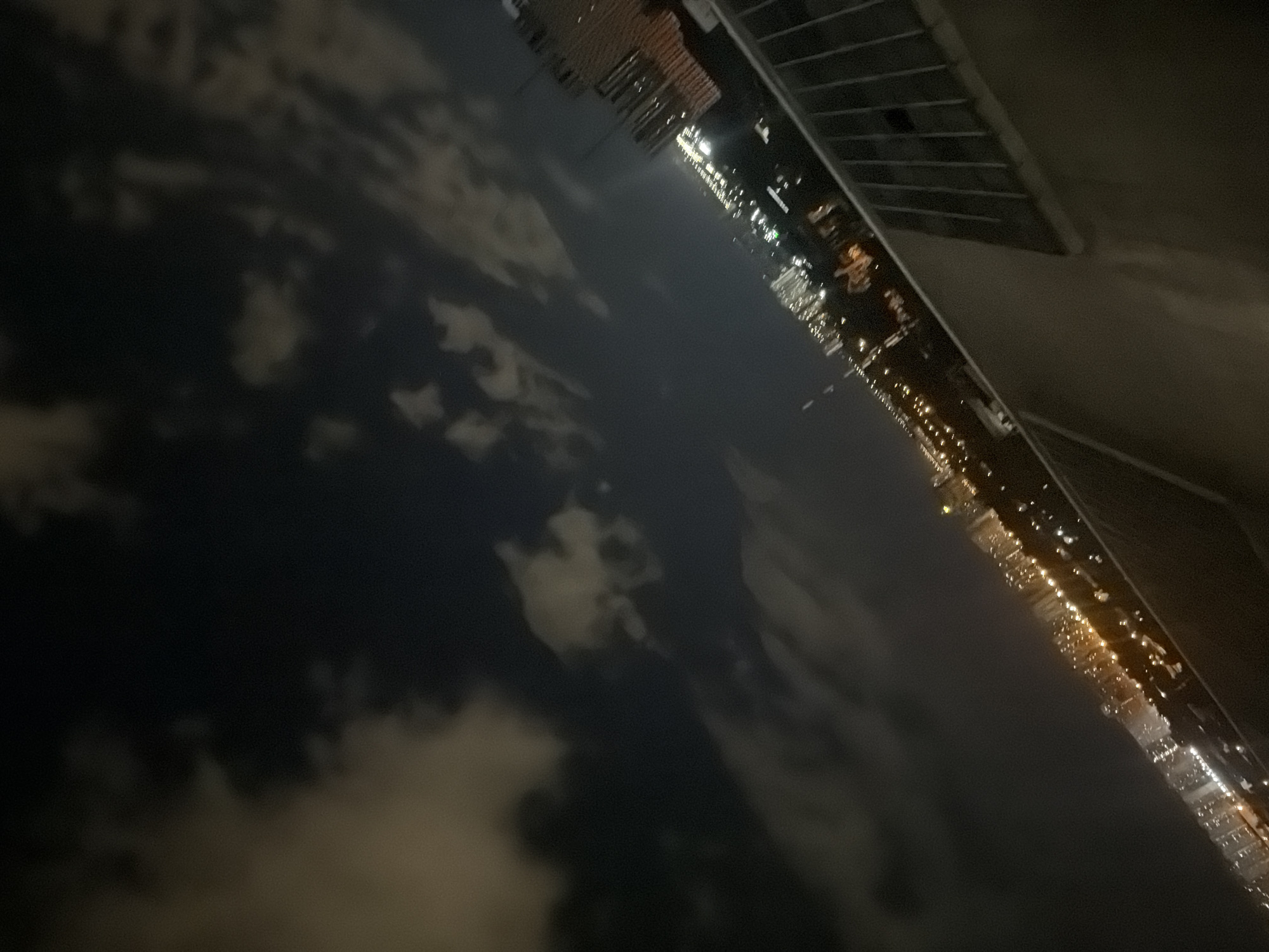 The image is a night-time, angled photograph taken from a high vantage point overlooking a cityscape. The sky is dark with scattered clouds, some illuminated from below by city lights. The city itself is visible as a sprawl of lights, suggesting densely populated areas with buildings and infrastructure. The lights vary in color, with some appearing white and others warm orange or yellow, indicating different types of illumination and structures. In the foreground, there is a railing, suggesting the viewpoint is from a balcony or rooftop. The overall impression is one of a vibrant, active city at night, viewed from above. The image does not contain any identifiable individuals or specific landmarks that would allow for precise identification of the location within Kyiv, Ukraine.