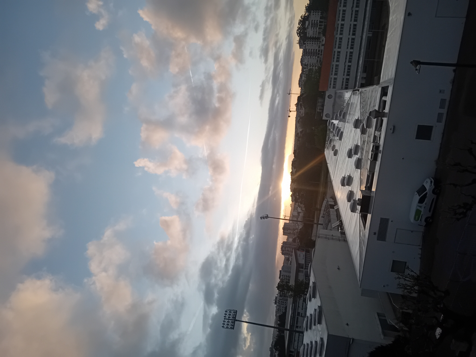 The image shows a cityscape at dusk. In the foreground, there's a large, flat roof with ventilation ducts. Next to it is a white van and a building with a light pole in front. In the midground, there are buildings and streetlights. In the background, the sky is filled with clouds. A sunset is visible on the horizon between the buildings. There is also a line in the sky, which appears to be the contrail from a plane. There's no direct evidence of location other than the architectural style of the buildings and the vegetation.