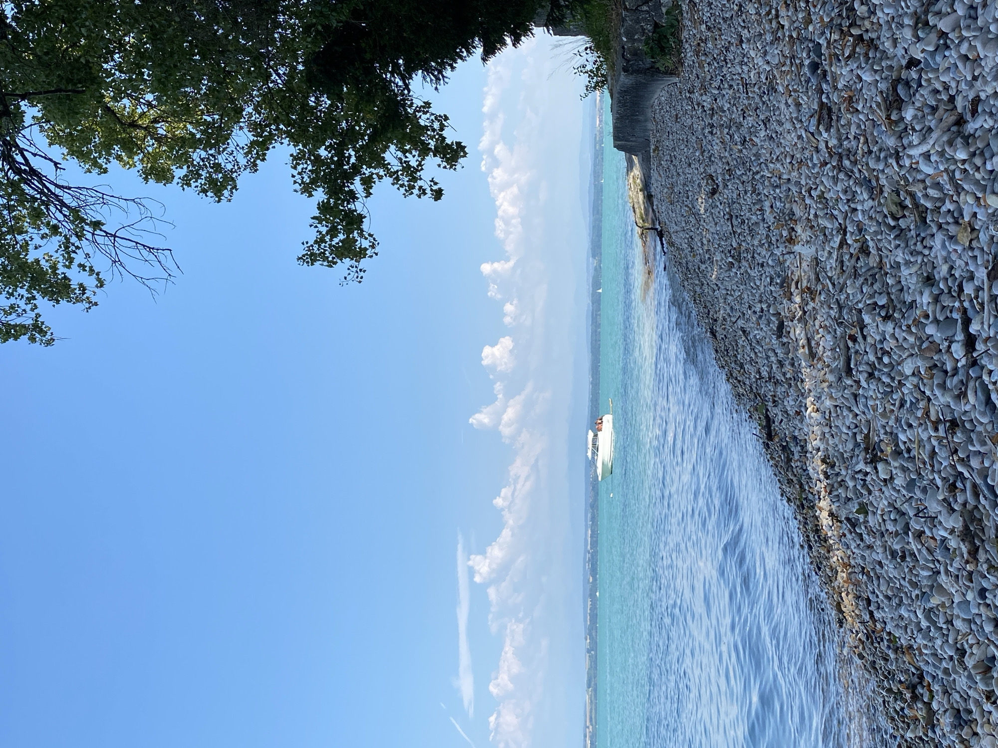 A wide-angle, vertical shot captures a serene lakefront scene with a stone-covered beach in the foreground and a body of water extending into the distance. The sky is a clear, vibrant blue with scattered fluffy white clouds. On the right side of the frame, the lush green foliage of a large tree drapes over the scene, casting shadows on the beach. A low stone wall borders the edge of the beach.
In the mid-ground, a small white motorboat with an open cabin is visible on the turquoise water, appearing to be at rest or moving slowly. In the far distance, a hazy, blue-toned landmass with rolling hills is visible on the horizon, suggesting a distant shoreline or mountains. The water is calm with gentle ripples. The beach is densely packed with smooth, gray and white pebbles, interspersed with some dried leaves and small pieces of driftwood. The overall atmosphere is peaceful and idyllic, evoking a sense of summer tranquility. The image does not contain any people. The scene is located in the city of Cortaillod, Switzerland.