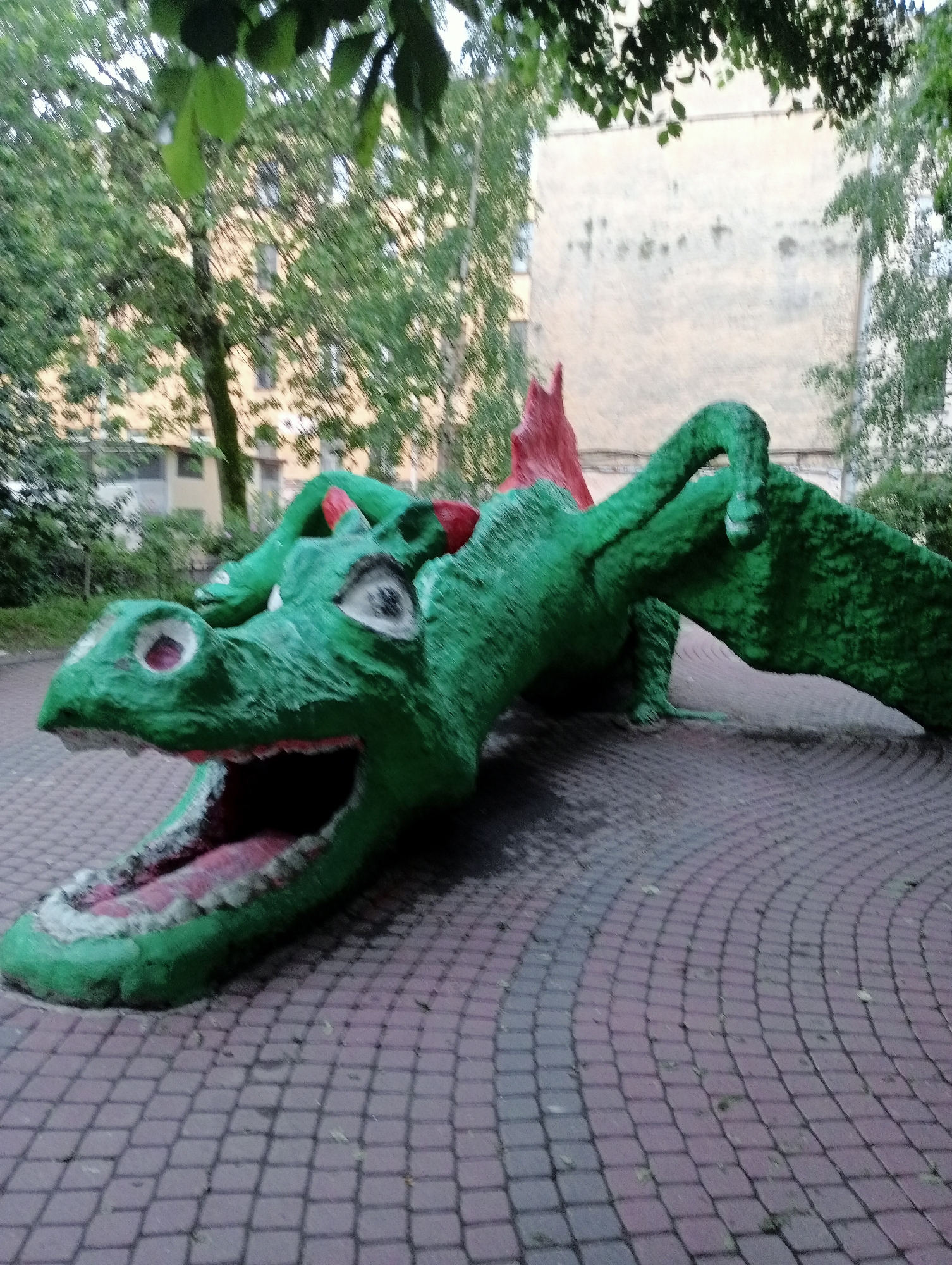 The image shows a vibrant green sculpture of a dragon lying on a cobblestone path. The dragon has its mouth open, revealing pink and white teeth, and its eyes are wide open. Red accents can be seen on the dragon's head and back. The sculpture is positioned outdoors, with trees and a building in the background. The cobblestone pavement appears worn, with some leaves scattered on it. The overall scene suggests an urban park or playground setting in Saint Petersburg, Russia.