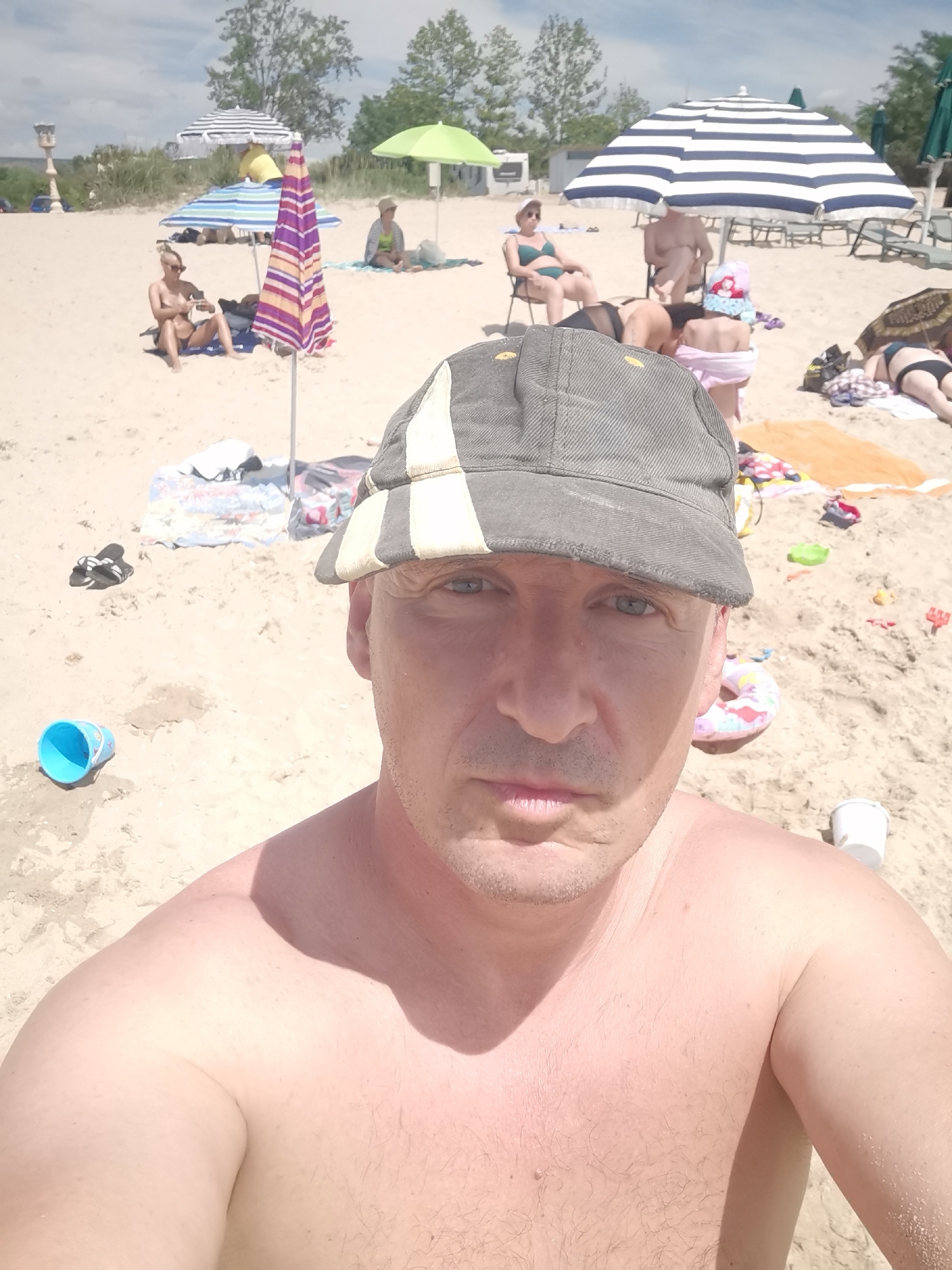 A man wearing a grey and beige baseball cap is taking a selfie on a sandy beach. His shoulders and upper chest are visible. In the background, several people are seen relaxing on the sand under umbrellas. Some are sitting in chairs, while others are lying down. There are beach umbrellas, towels, and beach toys scattered around the scene. The environment appears to be a sunny day with a partly cloudy sky, typical of a beach location in Kranevo, Bulgaria.