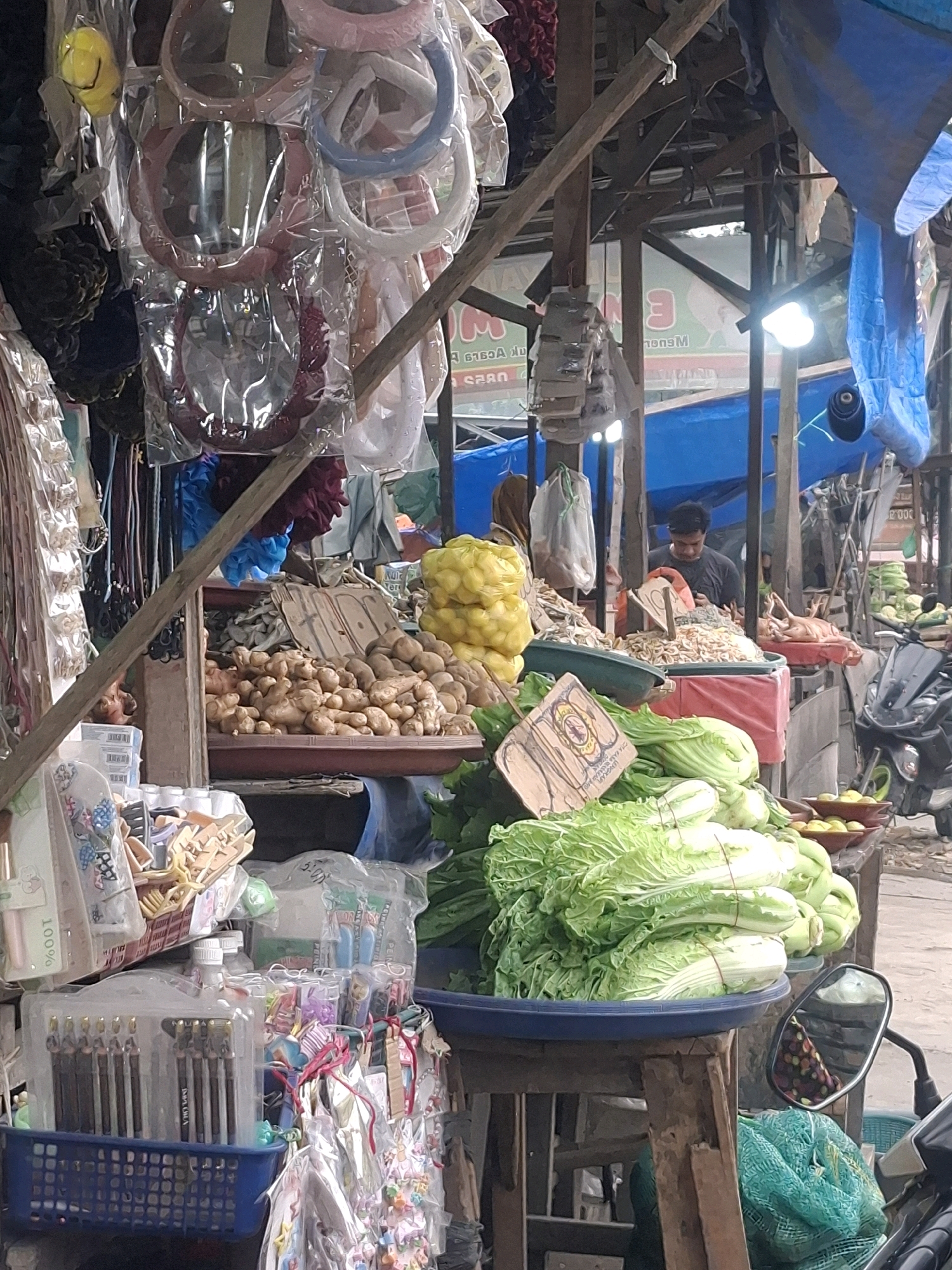 This is a medium shot of a market stall in Kenangan, Indonesia, likely a street market, with a focus on various merchandise. In the foreground, a large blue basin holds several heads of Napa cabbage, bound with red twine. To the right, a motorcycle mirror reflects a person wearing a patterned cloth. Behind the cabbage, various food items are visible, including a pile of ginger root, and yellow produce that appears to be some type of squash or gourd. Scattered among the produce are bags of small, dried fish or shrimp.
The stall structure is made of rough wooden beams and poles, with tarps and fabric serving as roofing and backdrops. A bright blue tarp is prominent in the upper right of the frame. Various goods are displayed, including hair accessories like headbands and clips, and packaged stationery items such as pens or pencils.
In the background, a man wearing a dark t-shirt is visible, appearing to be attending the stall and looking at his phone. The overall impression is of a bustling, possibly outdoor market setting with a variety of goods for sale. The lighting suggests it's daytime, with some artificial light sources, likely bulbs, also present.