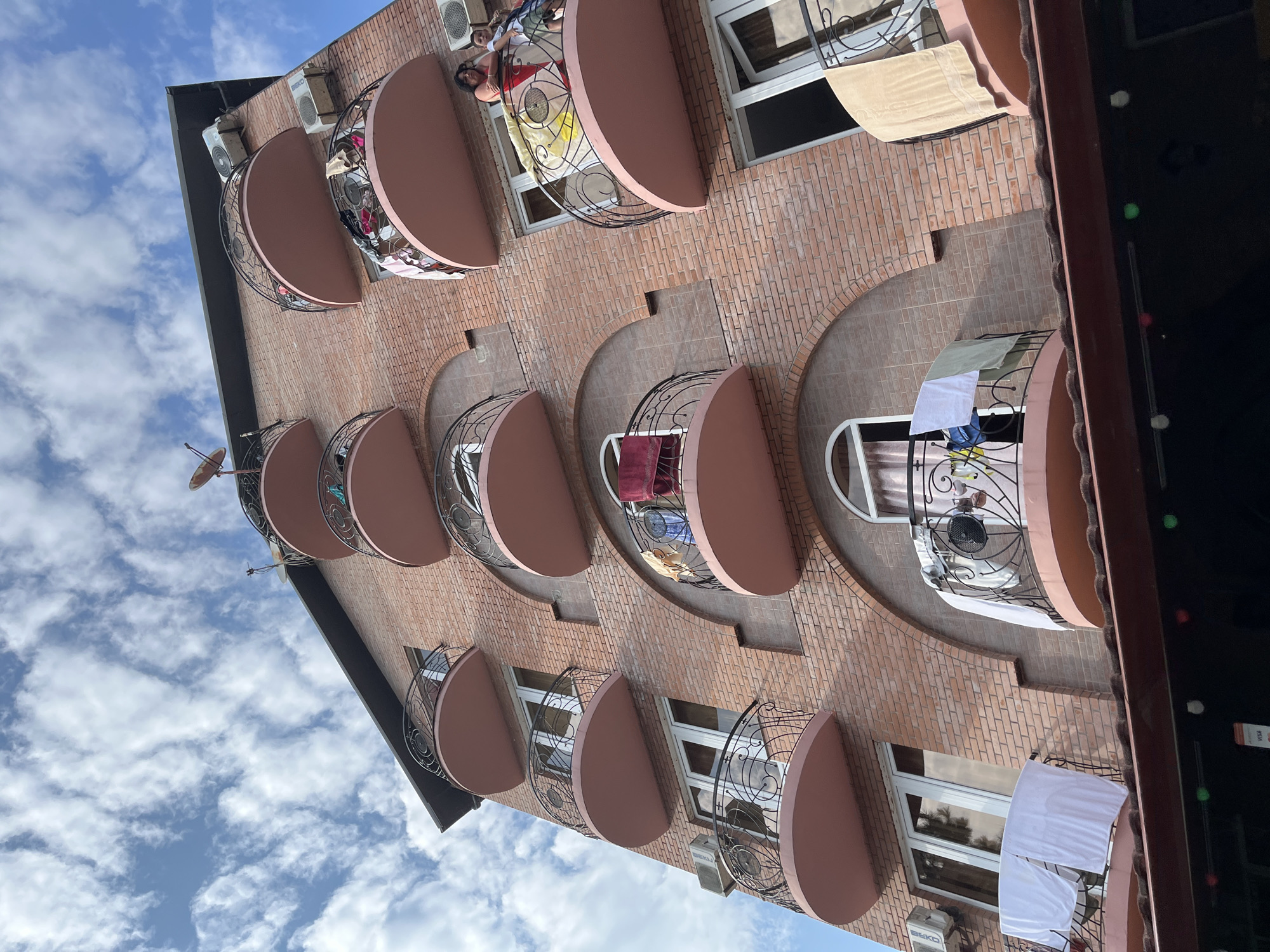 The image shows a multi-story brick building with rounded balconies on multiple levels. Laundry is hanging on the railings of several balconies, including white sheets, towels of various colors, and some clothing. Two women and a child are standing on one of the balconies. The building appears to be in a city or resort area, with the sky visible above and what looks like a darker covered area with lights at the bottom of the frame. The architecture and the presence of laundry hanging outside suggest a warm climate and possibly a residential or guesthouse setting. The building's design with arches above some windows and the protruding balconies is notable. The context provided indicates the location is Ureki, Georgia.