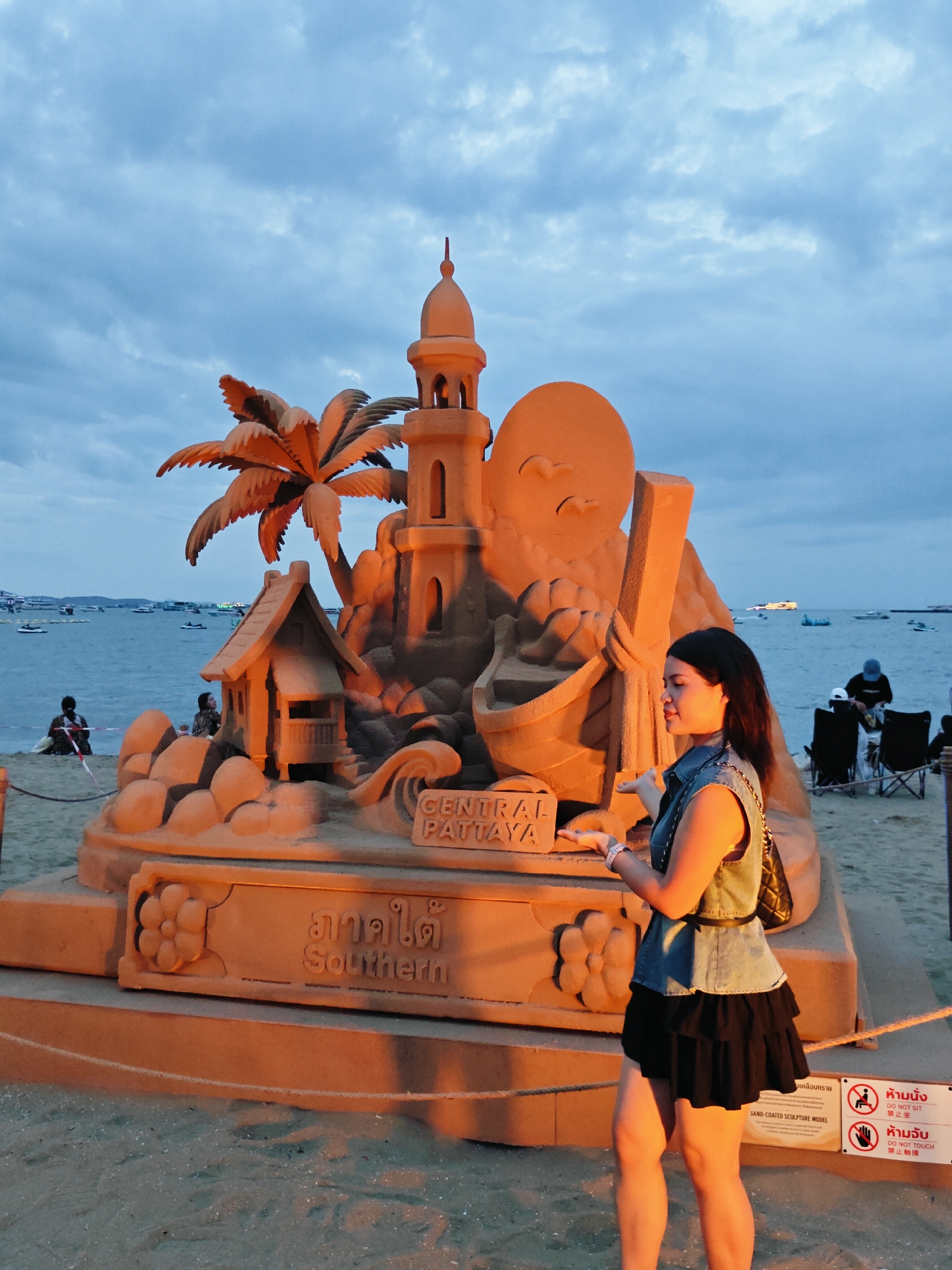 A woman stands on a sandy beach in Pattaya City, Thailand, posing with a large sand sculpture. The sculpture depicts a tropical scene with a lighthouse, palm tree, a small house, and a boat. Text on the sculpture reads "CENTRAL PATTAYA" and "Southern". The woman is wearing a denim vest and a black skirt. In the background, the ocean is visible with several boats, and the sky is cloudy. There are also signs with instructions in Thai and English, indicating "DO NOT SIT" and "DO NOT TOUCH".