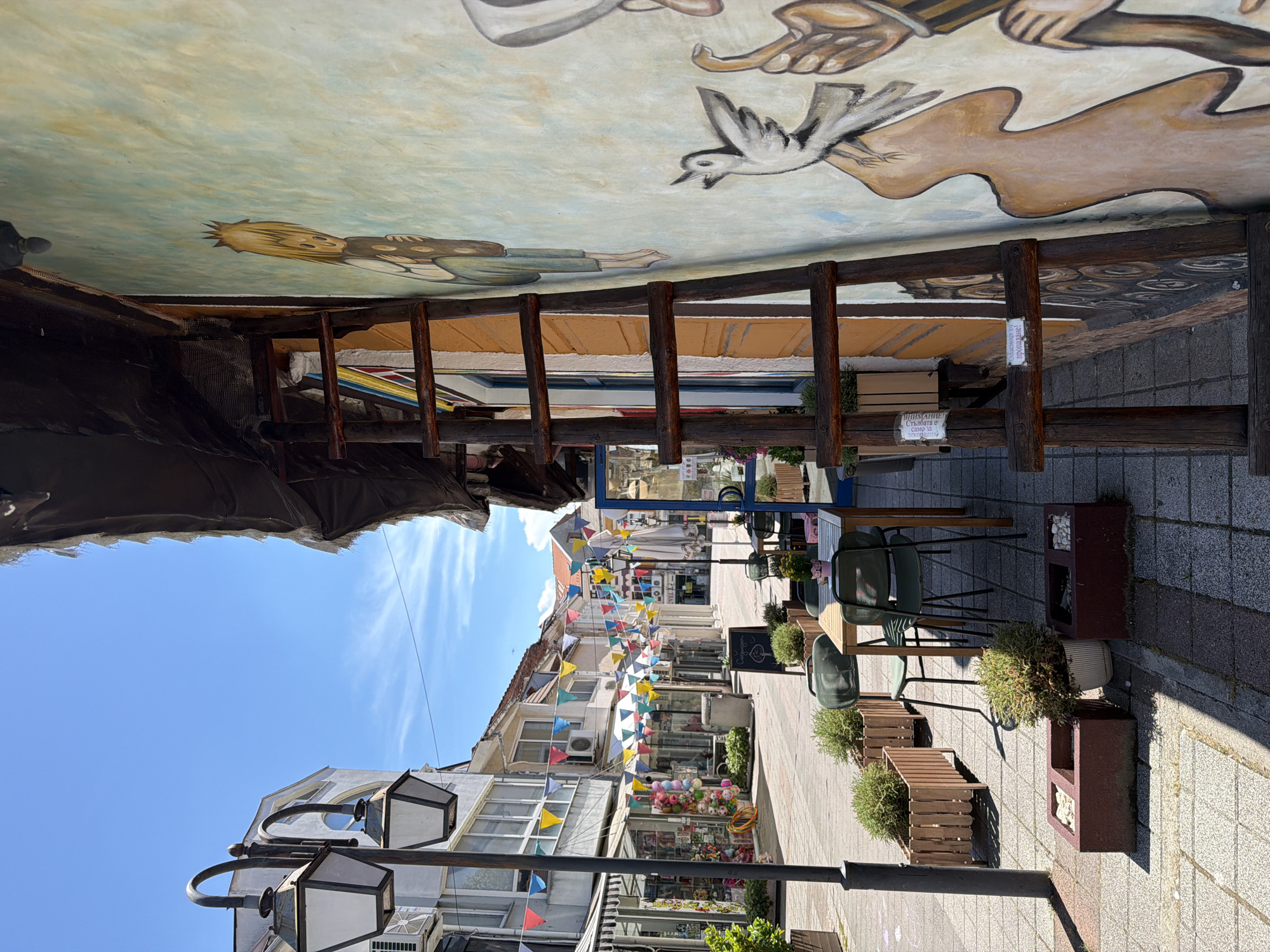 The image captures a street scene in Pazardzhik, Bulgaria, with a focus on the architecture and decorations. The perspective is from street level, looking upwards at buildings and a painted ceiling. The scene is illuminated by bright daylight, suggesting it's daytime.
Dominating the top portion of the image is a mural painted on what appears to be a ceiling or an overhead structure. The mural depicts stylized figures and imagery, including a person with blonde hair, a bird in flight, and abstract shapes. Below this, a rustic wooden structure, possibly part of an awning or a decorative element, extends into the frame.
The street itself is paved with stone tiles. Along the right side of the street, outdoor seating is arranged, with wooden tables and green chairs. Planters containing green bushes and small trees are placed along the walkway, adding a touch of nature to the urban environment. String lights with colorful triangular flags are hung across the street, creating a festive atmosphere.
The buildings lining the street are of varying architectural styles, some featuring balconies and windows. In the distance, more shops and facades can be seen. A prominent street lamp with multiple light fixtures stands on the left side of the image.
One visible sign, written in Cyrillic script, reads "ВНИМАНИЕ! Стълбите е само за посетители!" which translates to "ATTENTION! The stairs are for visitors only!" This suggests the presence of stairs nearby, possibly leading to another level or an establishment. The overall impression is a charming and lively street, likely a commercial or pedestrian area, decorated for an event or to enhance its aesthetic appeal. The weather appears clear and sunny, with a few wispy clouds in the sky.