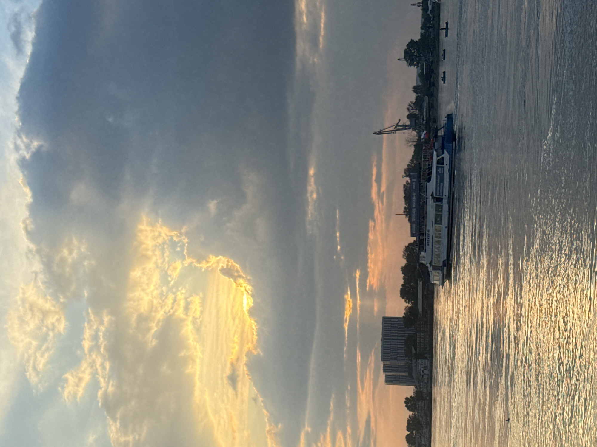A wide shot of the Angara River in Irkutsk, Russia, at sunset. The sky is filled with dramatic clouds, with the sun peeking through on the left, casting a golden glow and creating sunbeams. In the foreground, the river is calm, reflecting the colors of the sky. A white and blue passenger boat is moving across the water from right to left. In the distance, the silhouette of buildings and trees can be seen along the riverbank, as well as a large crane, indicating an industrial area. The overall scene is serene and picturesque.
