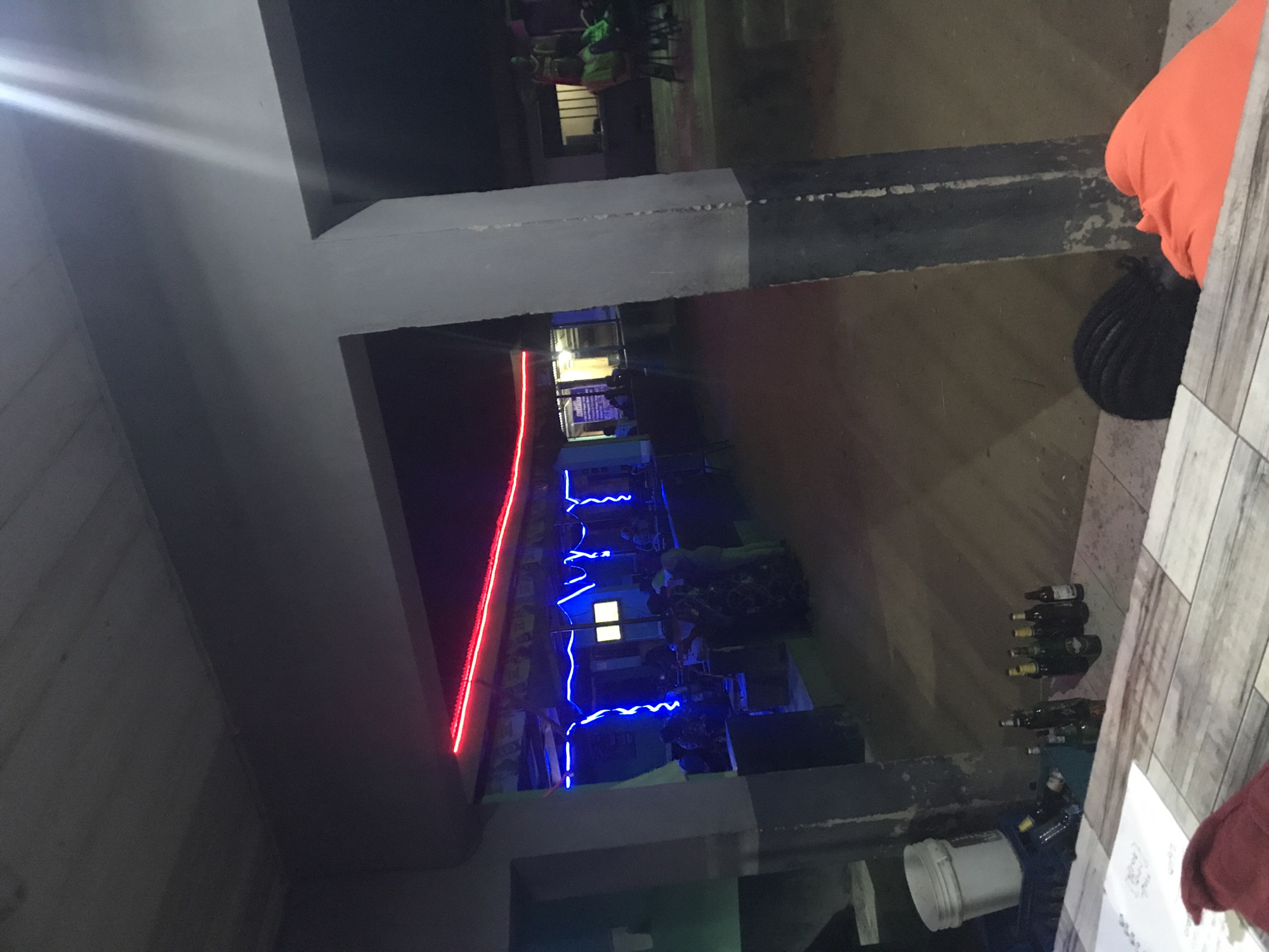 The image depicts an outdoor bar or restaurant in Kabong, Nigeria, during the night. A section of the establishment is visible from an elevated perspective. The space is illuminated with red and blue neon lights, and there are several patrons seated at tables. Some people appear to be standing near the entrance area. Near the right edge of the image, there are empty bottles on the ground, a white bucket, some documents, and part of a table. The structure above the space is made of concrete.