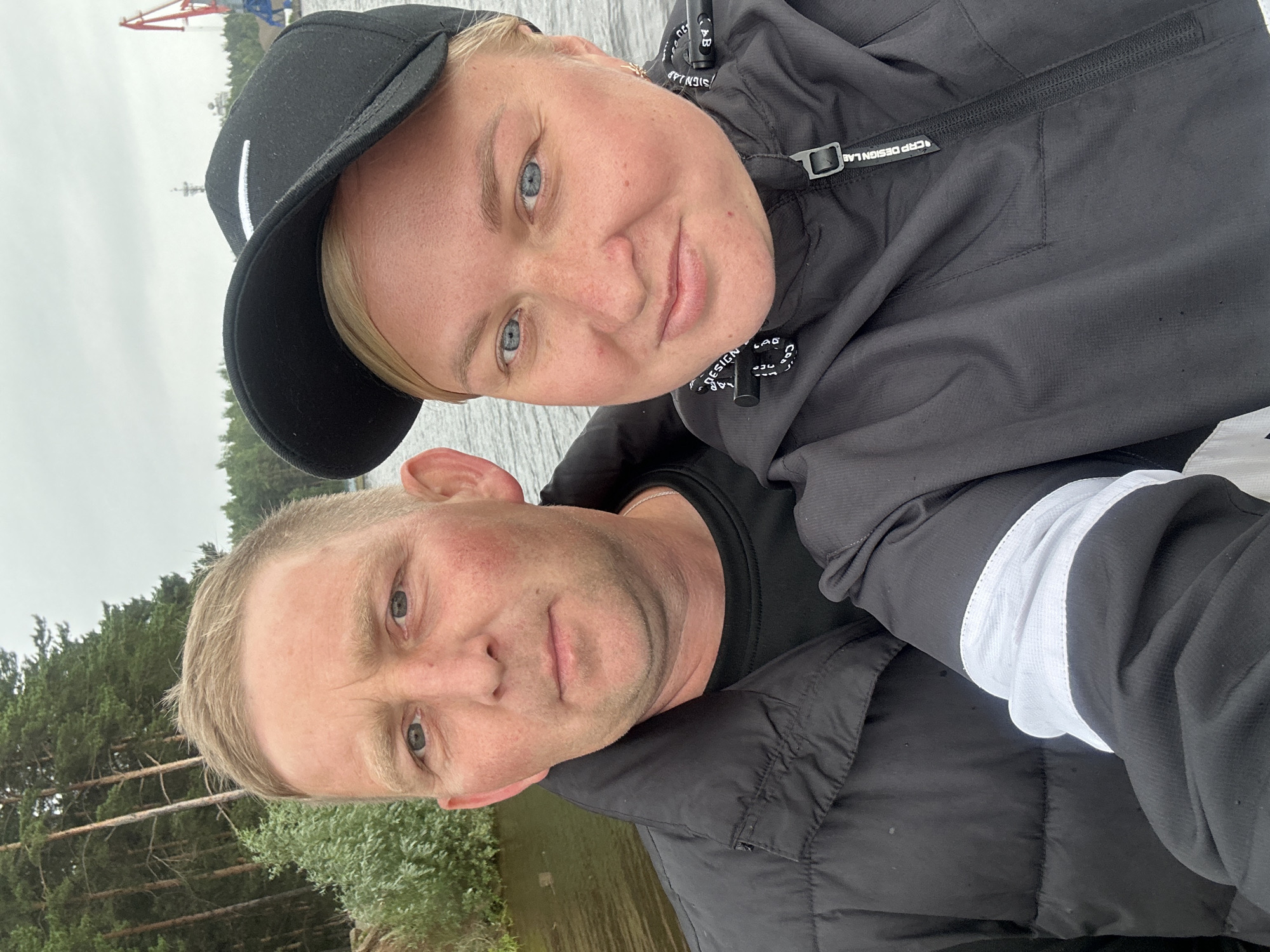 This is a selfie taken outdoors, featuring a Caucasian man and woman standing close together by a body of water. The man is on the left, wearing a black puffer jacket and a black t-shirt. He has short, light-colored hair and a fair complexion with some redness on his cheeks. The woman is on the right, wearing a black jacket with a white stripe detail on the sleeve and a black baseball cap. She has blonde hair and blue eyes, with a smattering of freckles across her nose and cheeks. The background shows a treeline with tall pine trees and a cloudy sky. In the distance, on the right side of the image, industrial cranes are visible, suggesting a port or industrial area near a natural setting. This scene is likely located in the city of Chaikovsky, Russia, given the specific industrial elements and the surrounding natural landscape often found in that region.