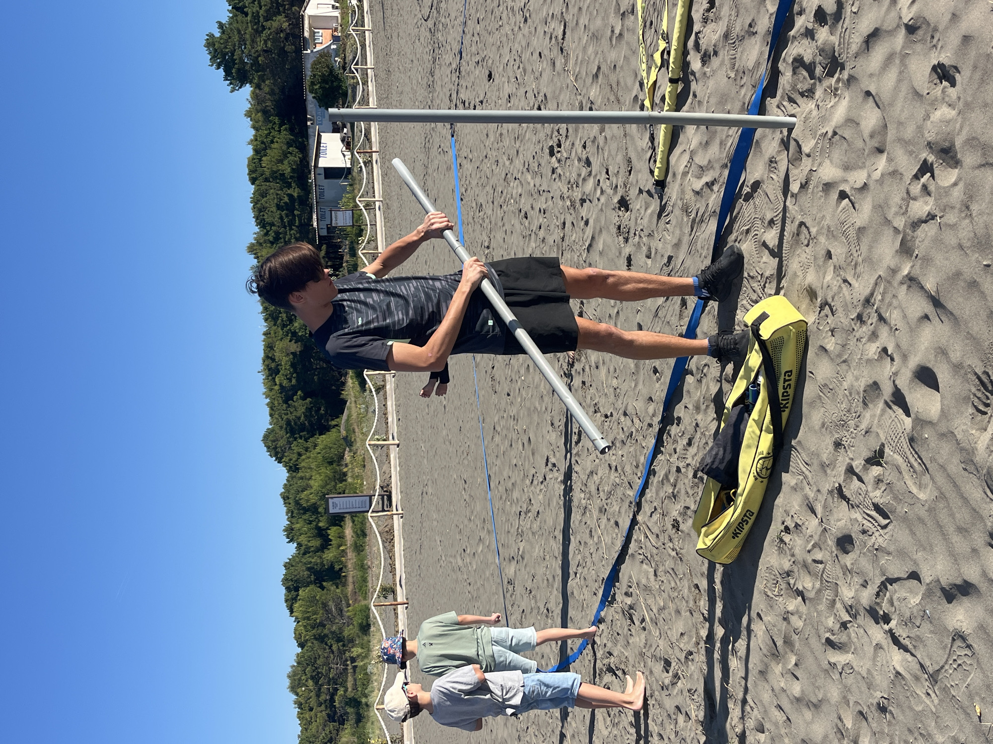 In Pistula, Montenegro, on a sunny day, a group of four individuals are gathered on a sandy beach, likely preparing for a recreational activity. The scene is set with a clear blue sky and a line of trees bordering the distant background, suggesting a coastal or park-like environment.
In the foreground, a young man with dark hair, wearing a black t-shirt with a geometric pattern and black shorts, is actively engaged in assembling what appears to be a piece of sports equipment. He is holding two sections of a gray pole, one in each hand, and is in the process of connecting them. His focus is on the task at hand.
To the left of the frame, two younger individuals are standing and observing. One, wearing a light blue t-shirt, light-colored shorts, and a beige baseball cap, faces away from the camera. Beside him, another young person, in a green t-shirt and light blue shorts, also looks towards the activity. Their postures suggest casual observation.
Further to the left, partially visible, is another person in a light blue t-shirt and light-colored shorts.
Scattered on the sand are various pieces of equipment. A yellow Kipsta bag, open and containing a dark item, lies on the ground. Also visible are yellow straps or bands and a blue line, possibly marking a boundary or part of the sports setup. A tall, gray pole stands upright in the sand, serving as an anchor point for the equipment.
The ground is sandy, marked with numerous footprints, indicating recent activity on the beach. In the mid-ground, a rope barrier with wooden posts is visible, separating the beach area from the treeline. Behind the barrier, a building with "TOILET" signs is situated, suggesting amenities are available nearby. The overall impression is of a relaxed, outdoor setting where a group is setting up for a game or activity.